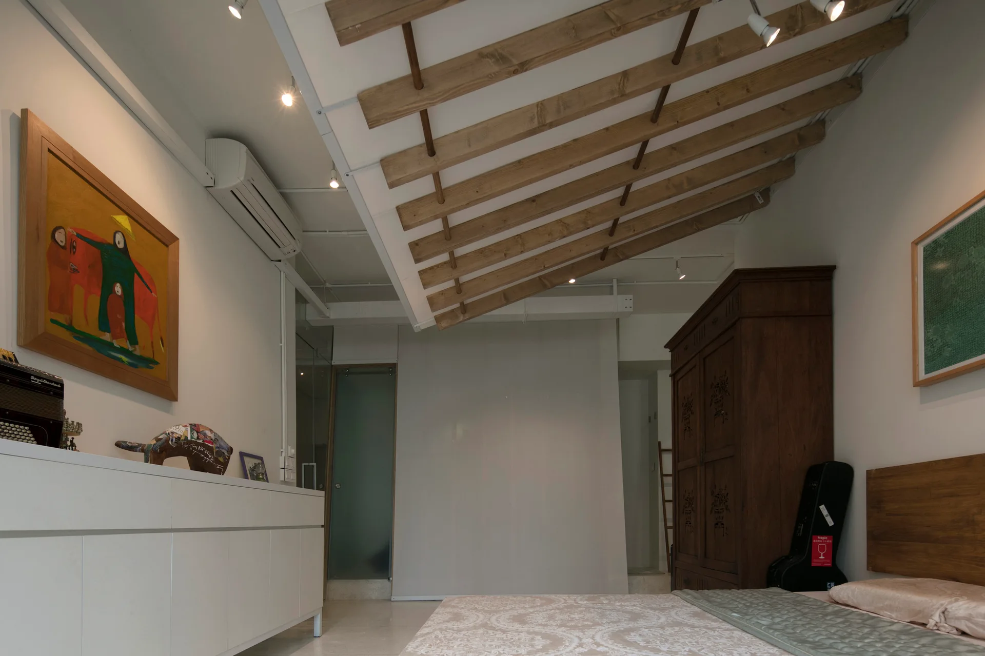 A modern transitional bedroom featuring exposed angled wood ceiling beams, a white media console, and a large wooden armoire.