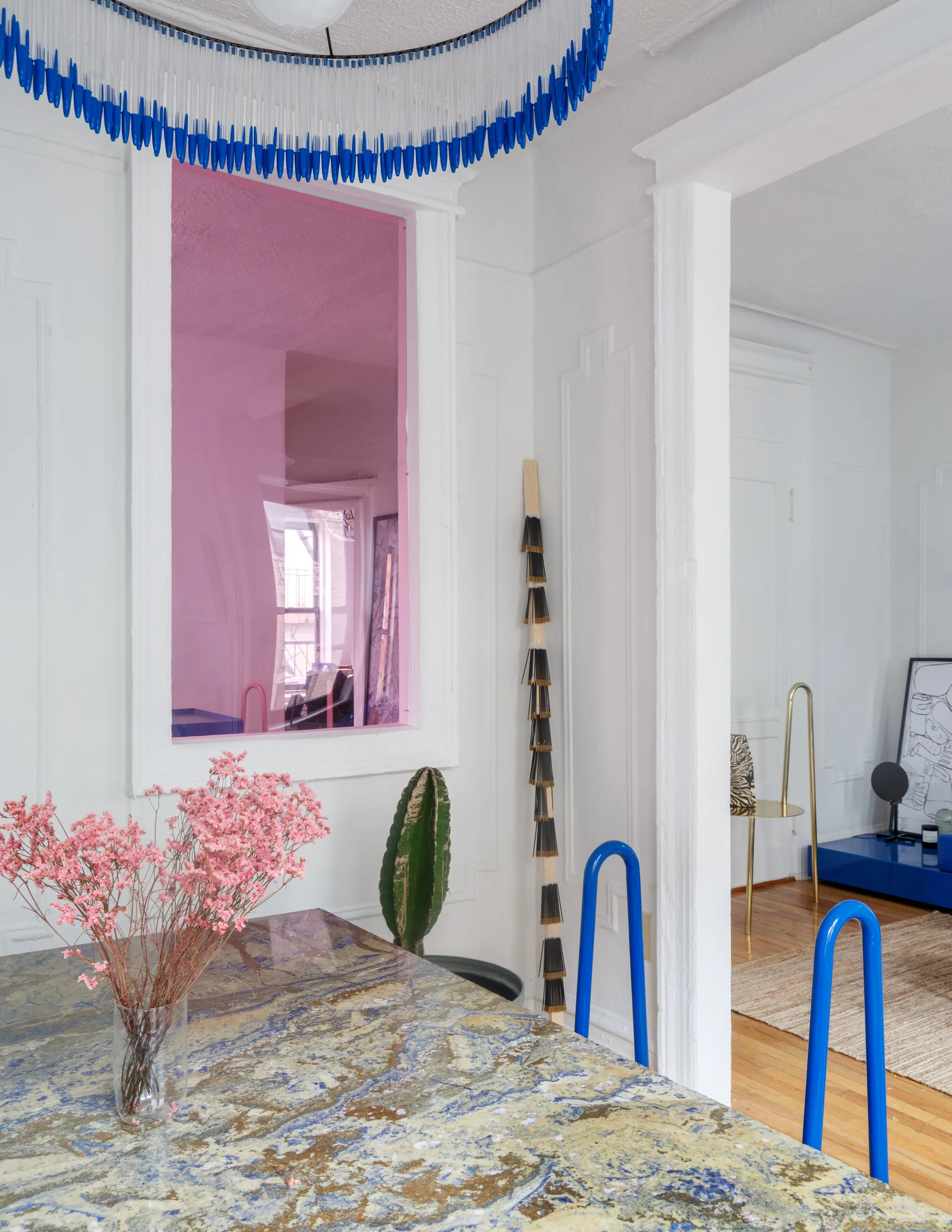 An eclectic dining room featuring a patterned marble table set beneath a blue fringe chandelier next to a pink glass interior window cutout.