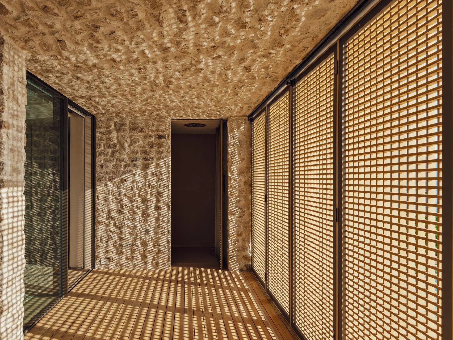 A transitional corridor featuring rough stone walls and ceiling, wood plank flooring, and large metal lattice screens that filter sunlight.