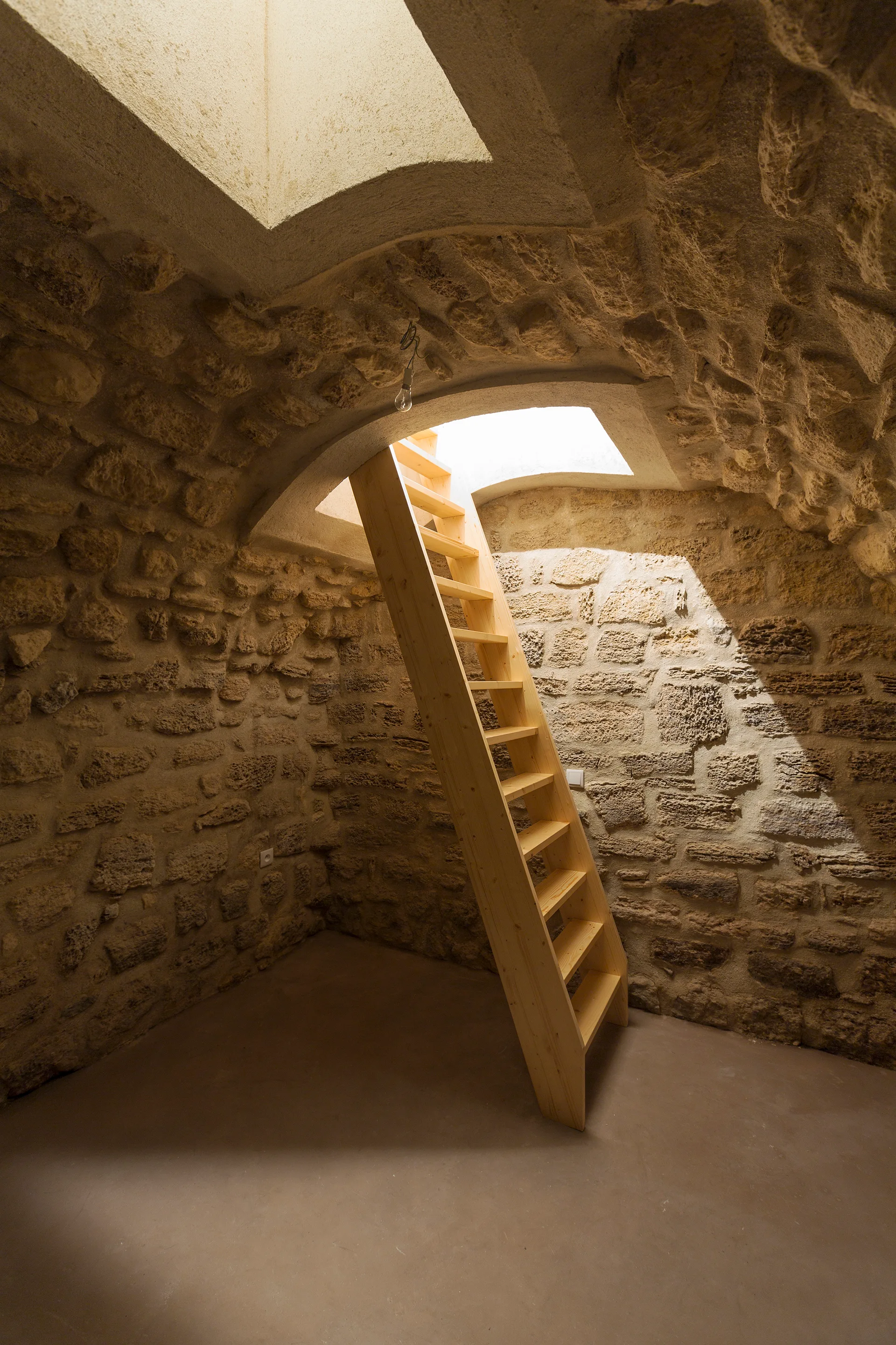 A simple wooden ladder staircase stands in an arched stone cellar, illuminated by bright natural light from a square ceiling skylight.