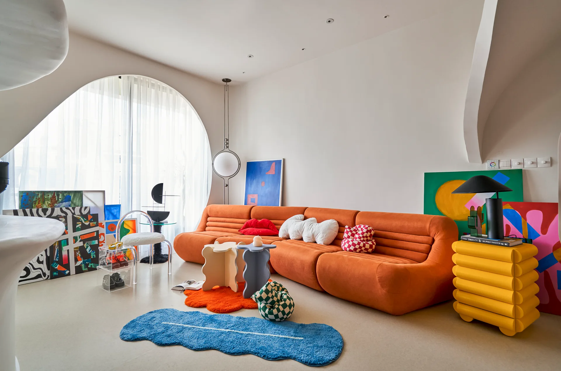 A postmodern living room features a long orange sofa, a large circular window with sheer curtains, colorful abstract paintings, and shaped rugs.