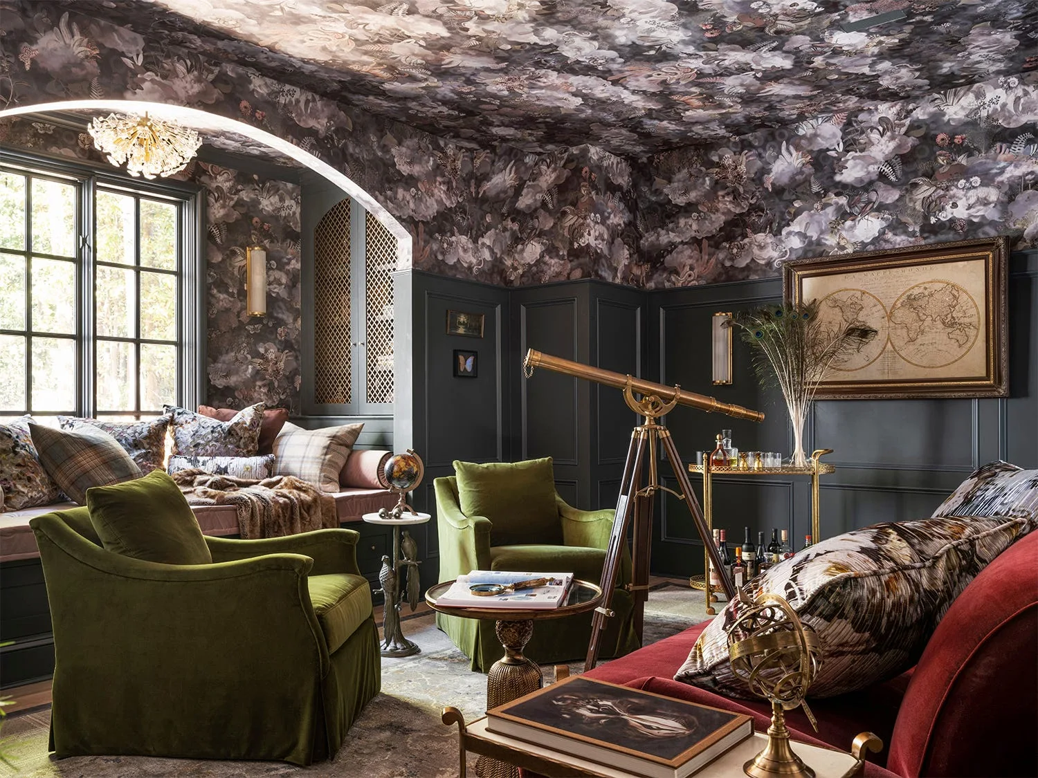 A moody library features dark wainscoting, floral wallpaper on the walls and ceiling, green velvet armchairs, and a brass telescope on a tripod.