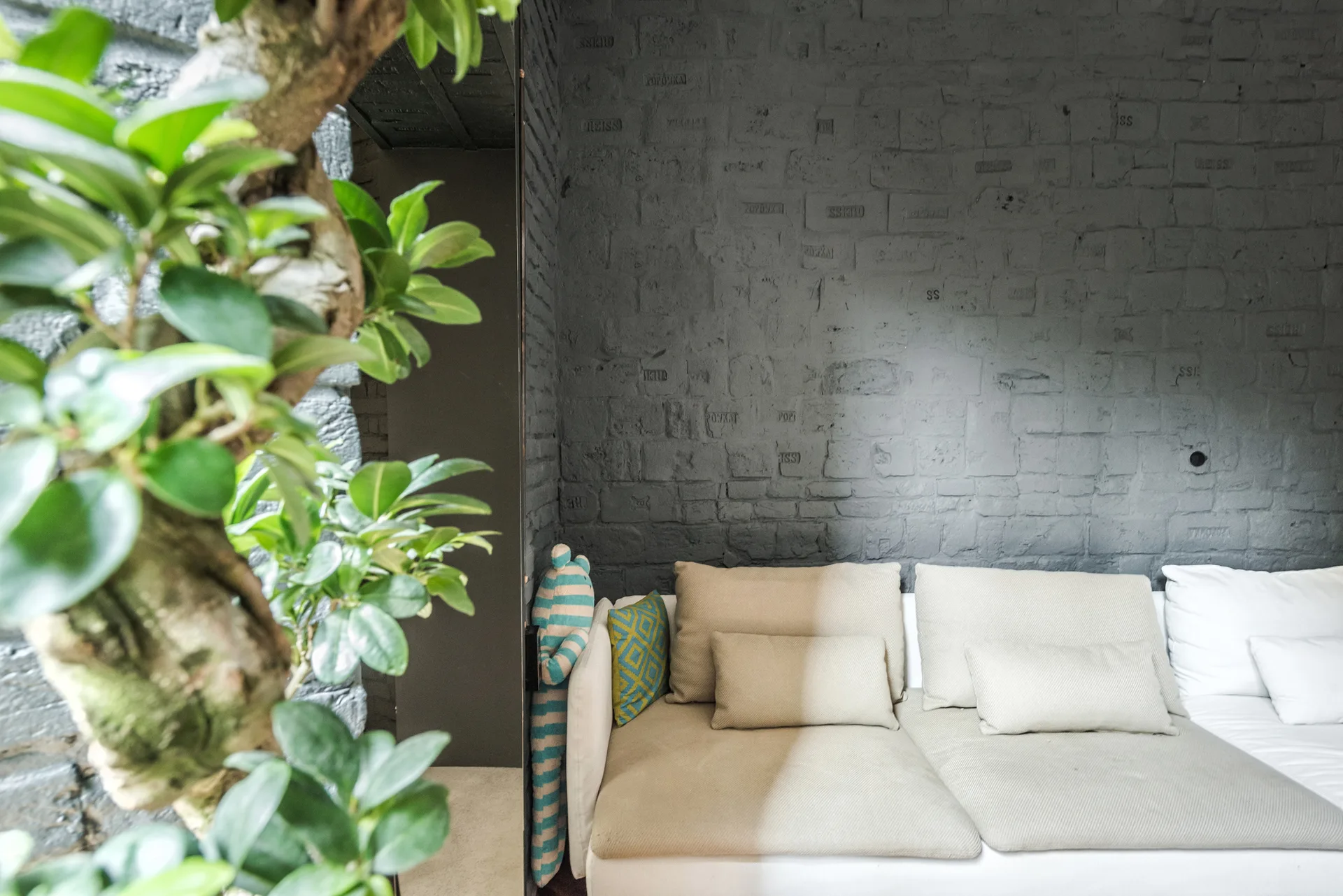 A beige sectional sofa with throw pillows is placed against a textured grey painted brick wall with green plant leaves in the foreground.