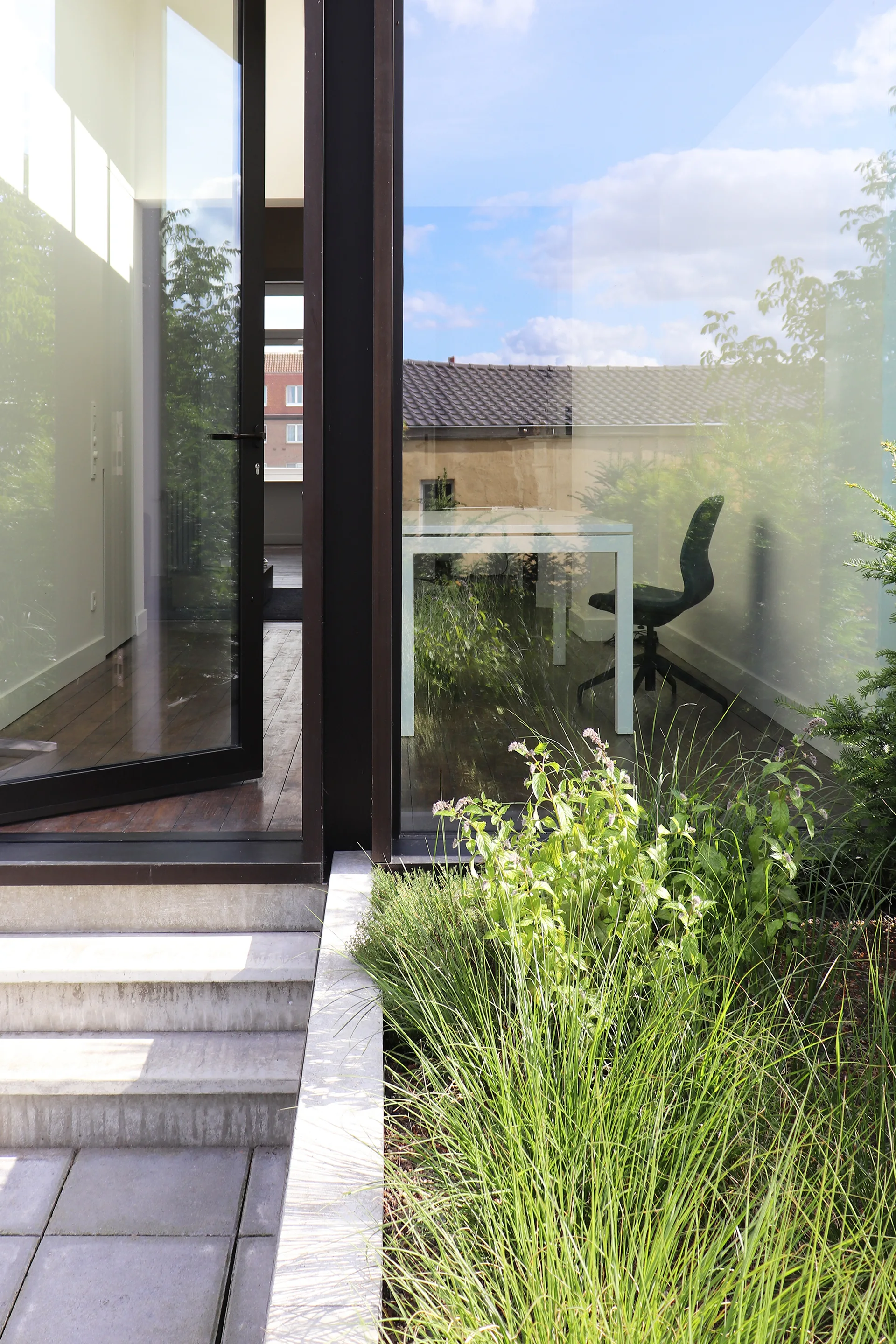 A view from a concrete patio with green grass looking through a glass wall into a minimalist home office with a white desk and dark wood floors.