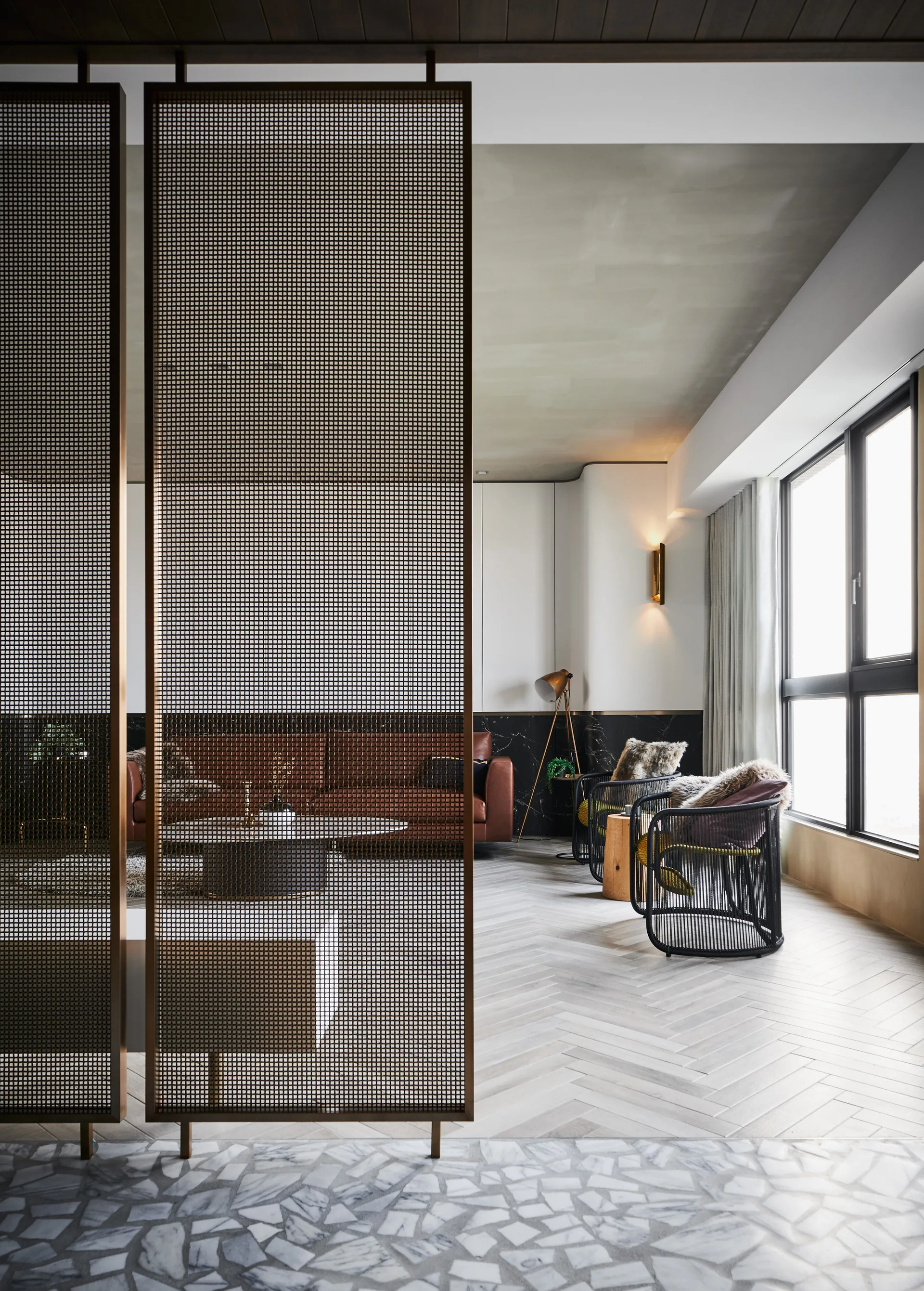 A modern living room features a bronze metal mesh divider, light wood herringbone flooring, a brown leather sofa, and two black corded lounge chairs.