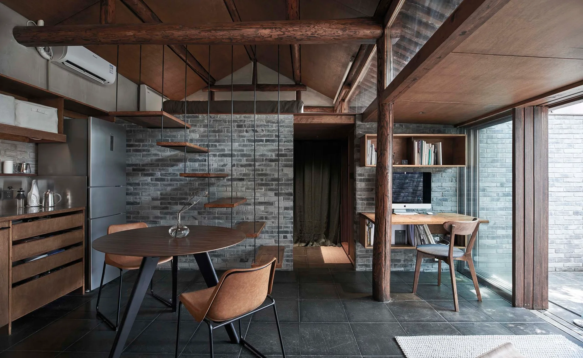 A compact residential space features a floating wooden staircase, a round dining table with tan chairs, and an office nook against a grey brick wall.