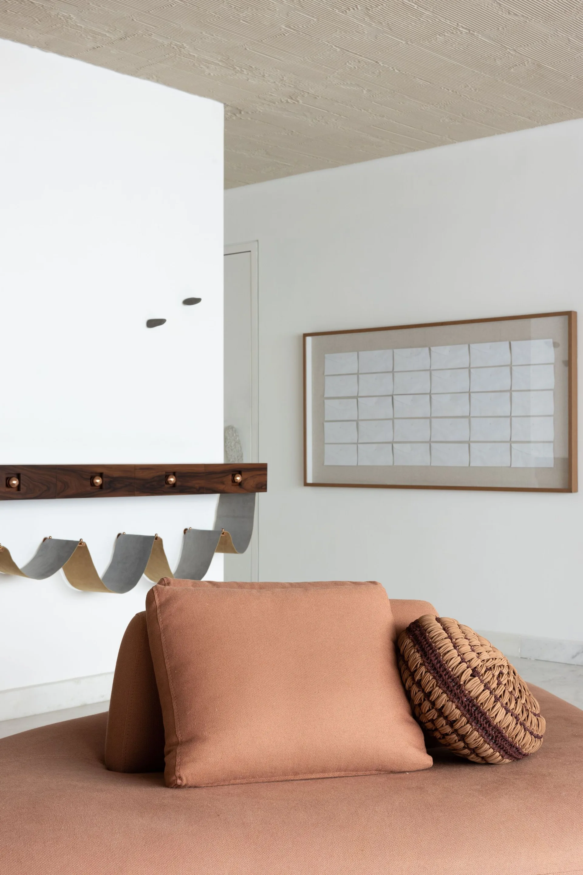 A close-up of a terracotta sofa cushion and a round woven pillow in front of a white wall featuring a wooden wall-mounted rack and a framed grid artwork.