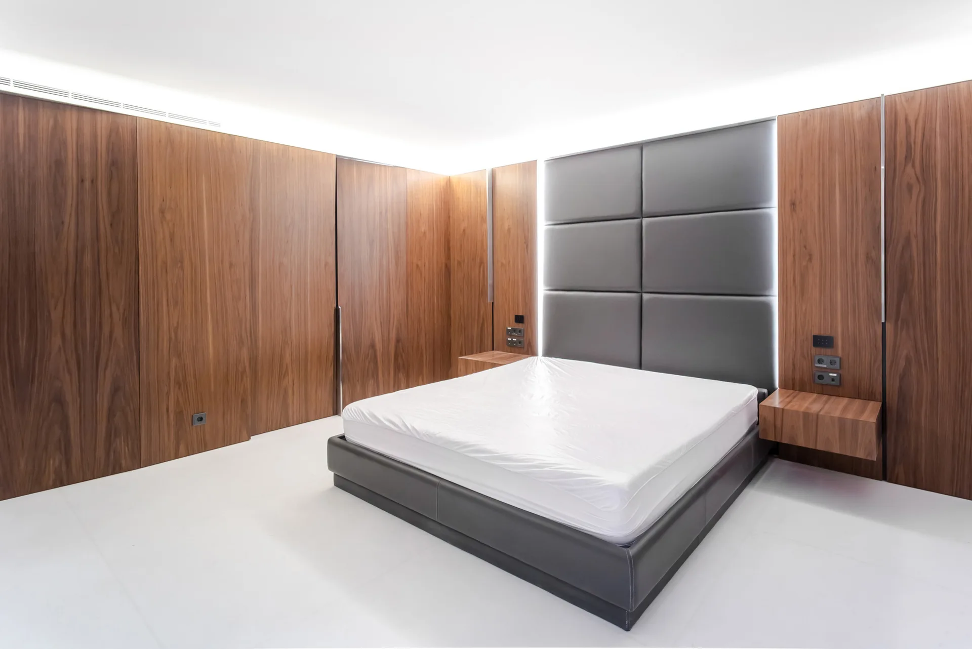 A modern bedroom featuring a grey upholstered bed and headboard flanked by wood-paneled walls with floating nightstands and cove lighting.