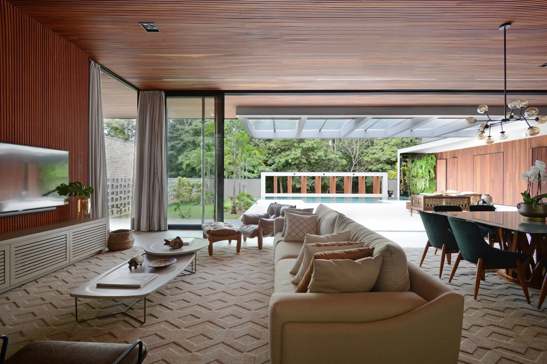 An open-concept living area with a wood slat ceiling features a cream-colored sofa, a geometric rug, and large sliding doors opening to a pool.