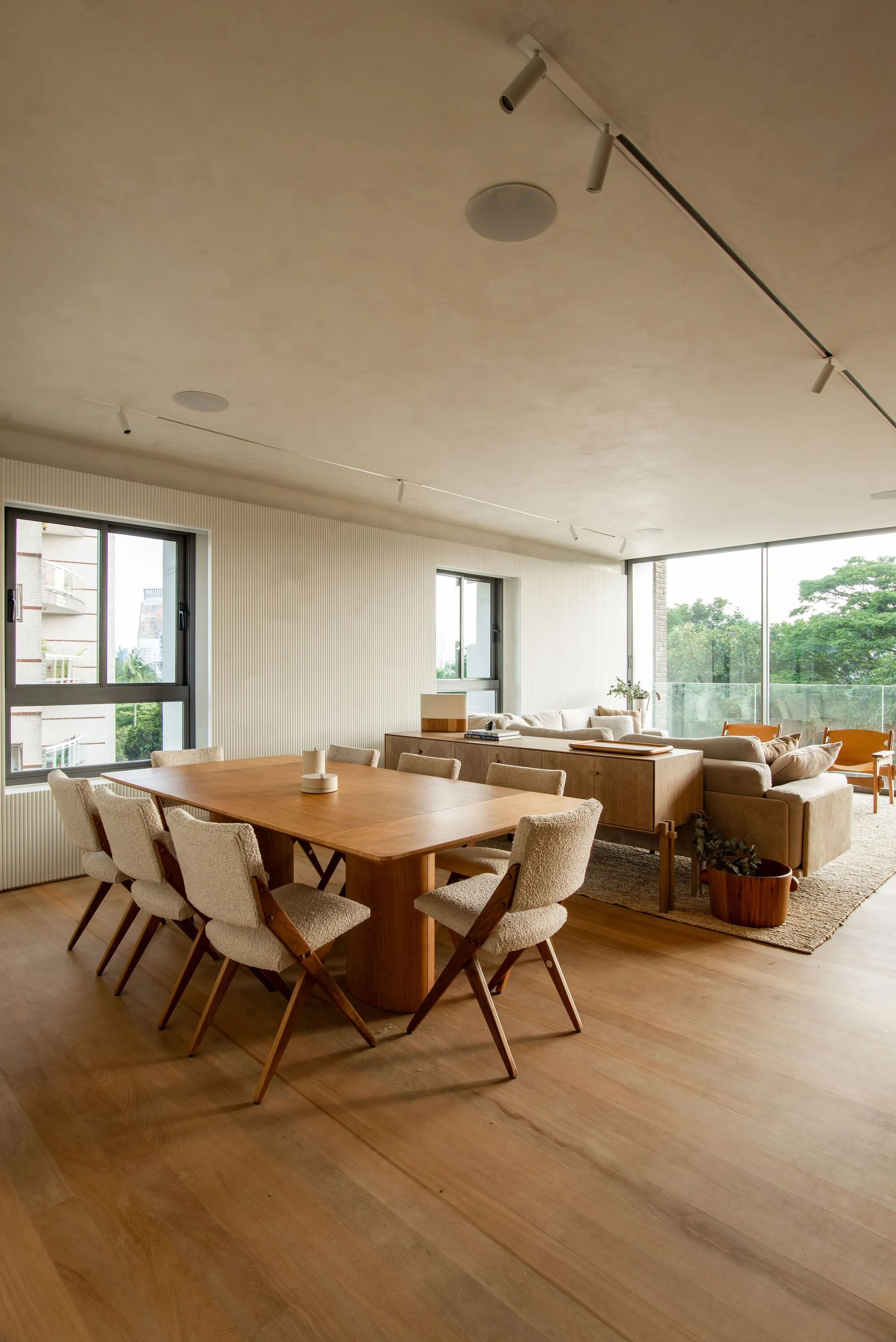 An open-plan dining area features a large rectangular wood table surrounded by cream bouclé chairs, set against a vertically fluted white wall.