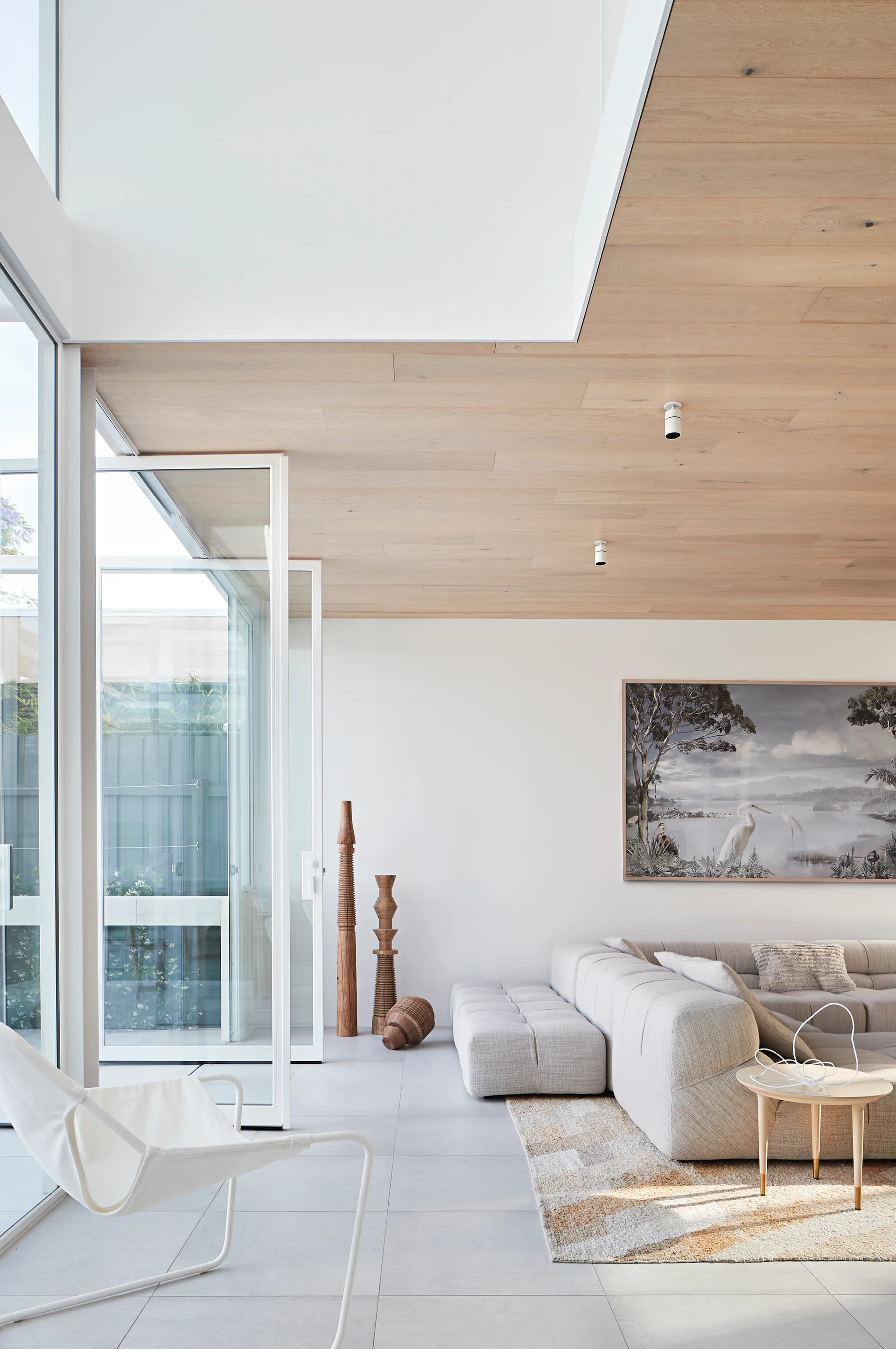 A bright living room features a modular grey sectional sofa, light wood ceiling planks, and tall glass doors opening to a courtyard.