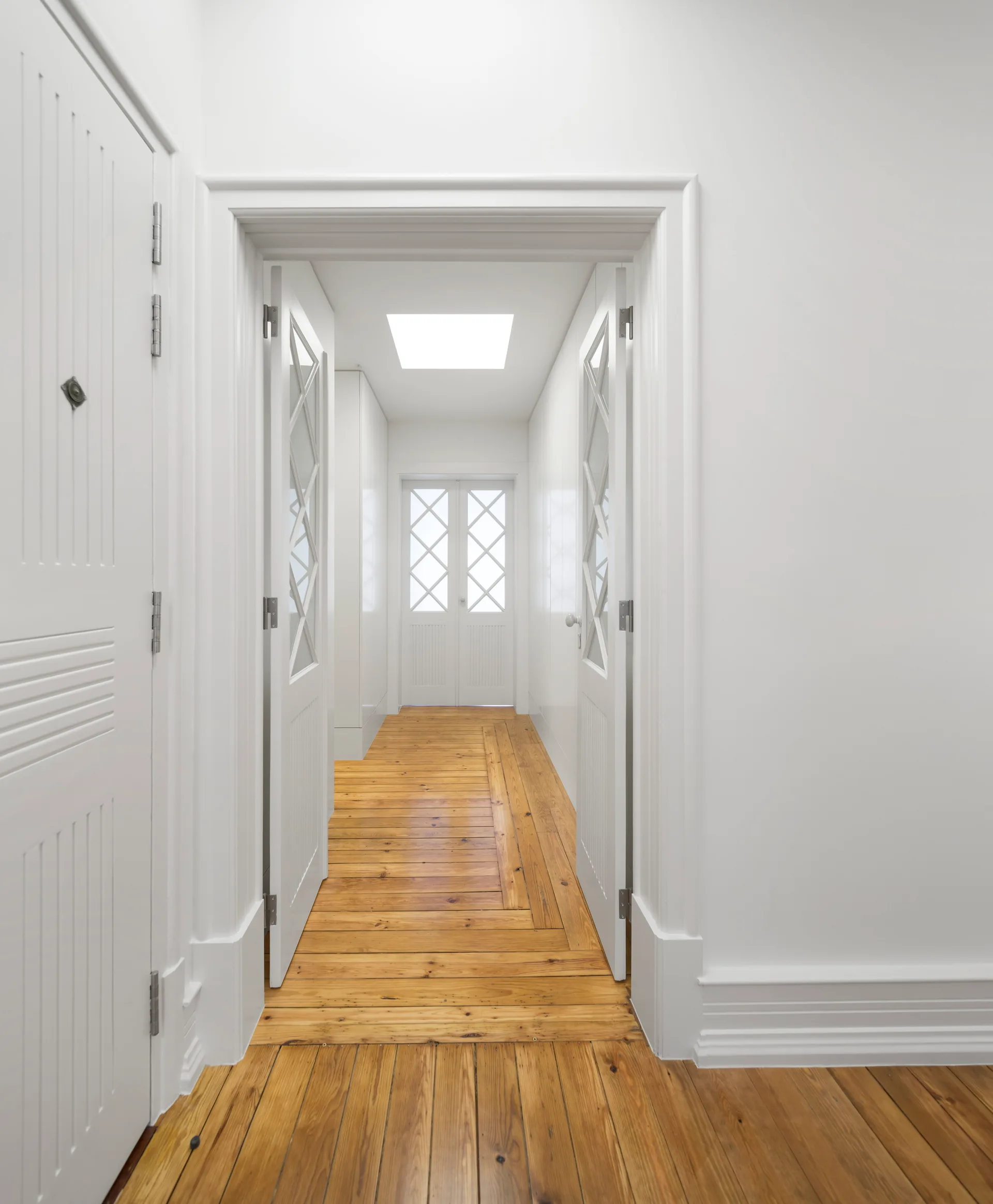 A bright white hallway features natural pine wood floors, white paneled doors with glass inserts, and a rectangular skylight in the ceiling.