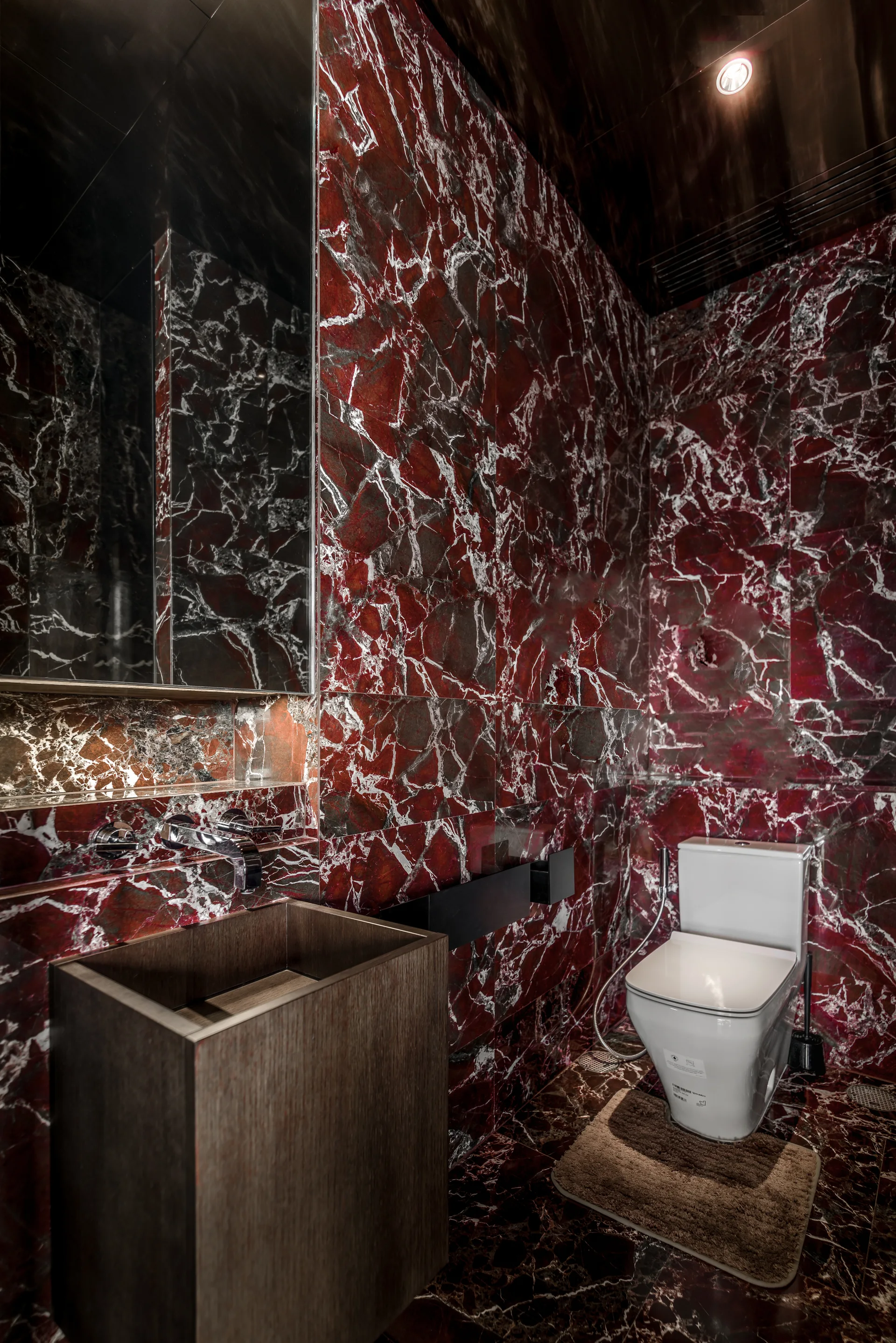 A modern powder room featuring polished red veined marble walls and floors, a blocky dark wood vanity, and a white toilet.
