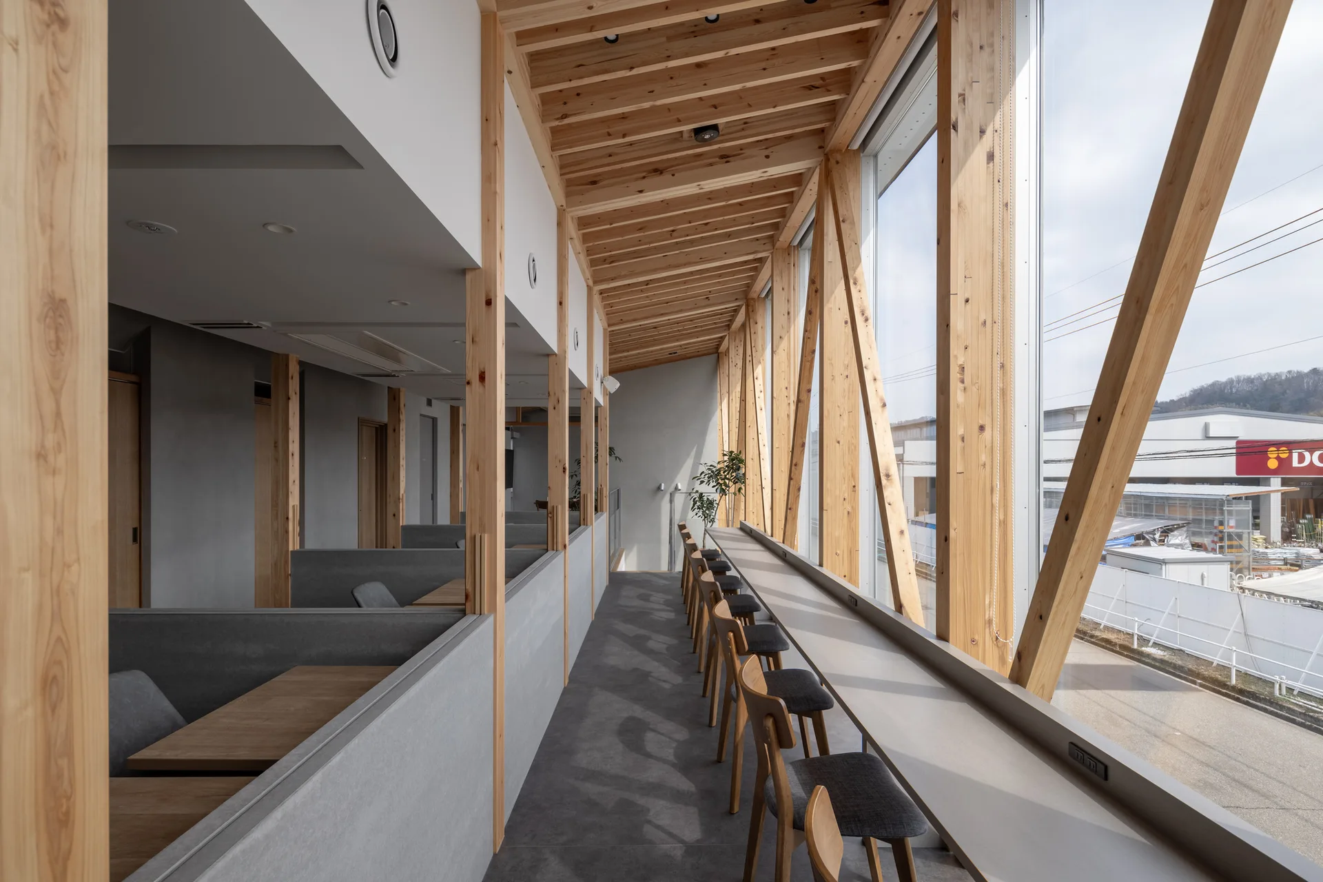 A modern workspace features a long grey counter with wooden chairs along a window wall beneath exposed timber framing and ceiling beams.