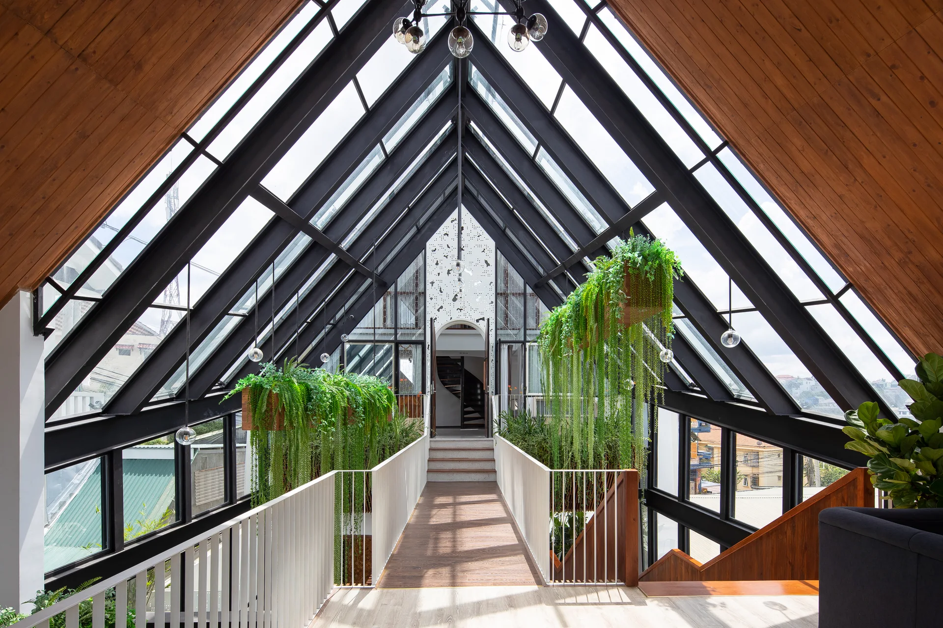 An elevated wooden walkway with white metal railings is framed by a black A-frame glass roof and features hanging planters with long green vines.