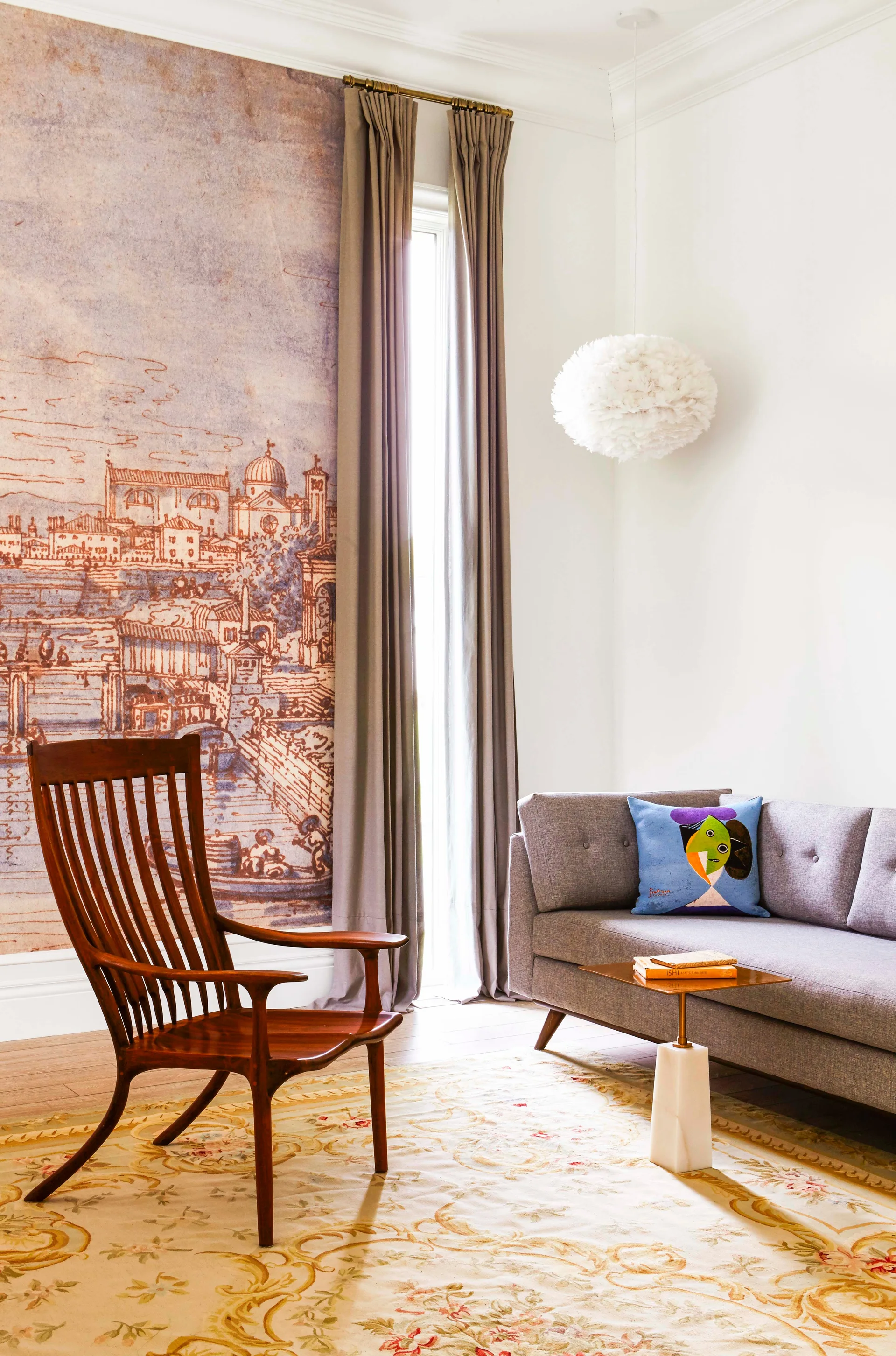 An eclectic living room features a grey sofa, a wooden slat-back armchair, and a large architectural wall mural next to tall grey curtains.