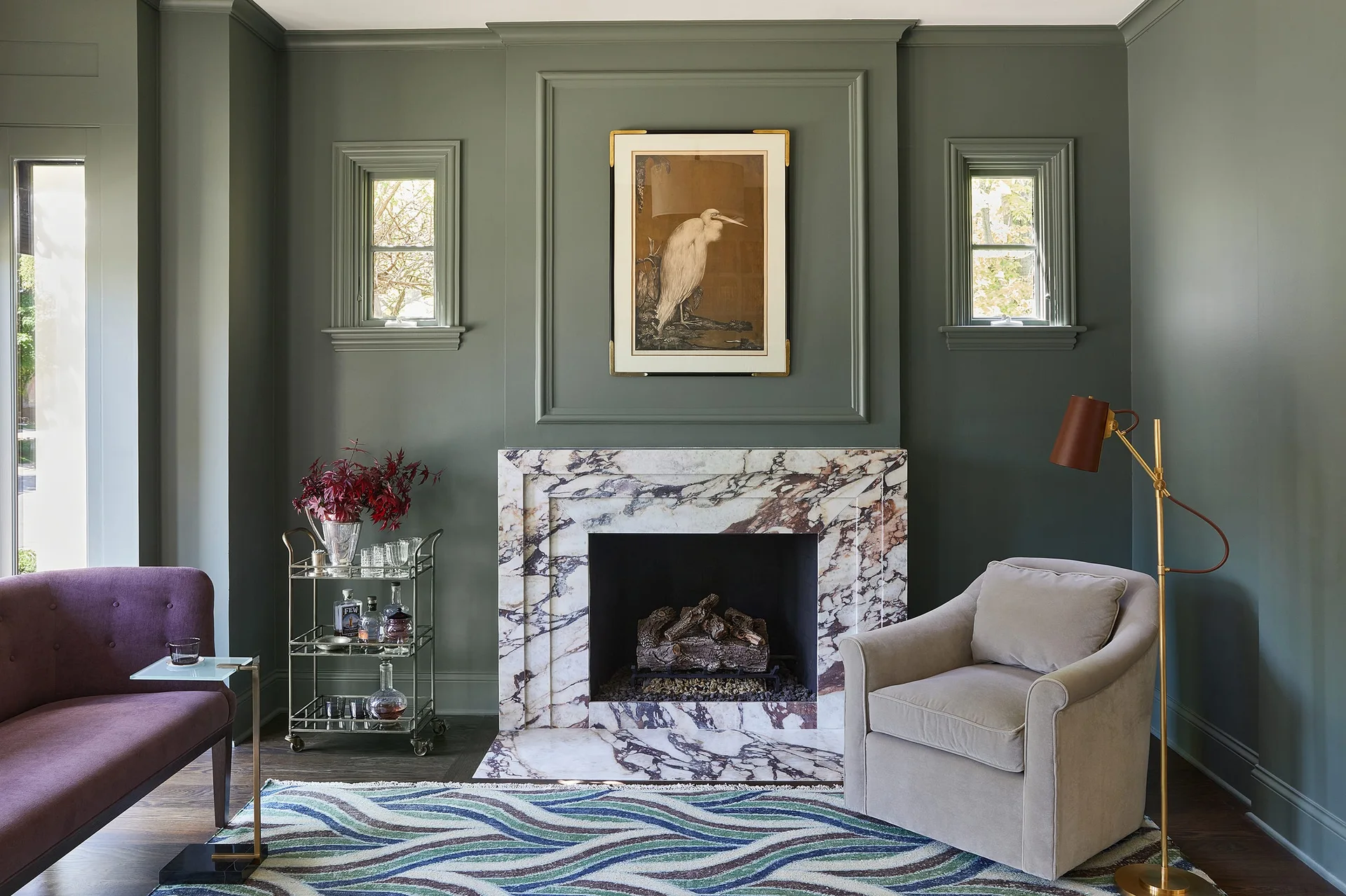 A living room featuring sage green paneled walls, a heavily veined purple and white marble fireplace, and a light gray armchair on a wavy rug.