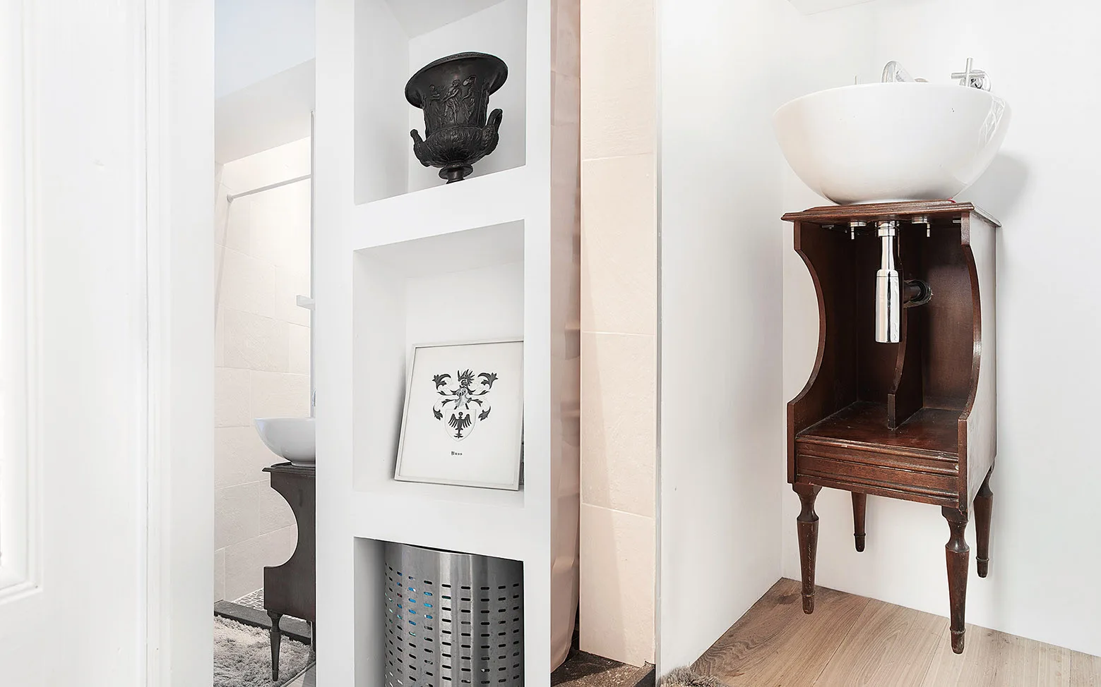 A white ceramic vessel sink sits atop a dark vintage wood washstand next to recessed white bathroom shelving containing a decorative urn and framed art.