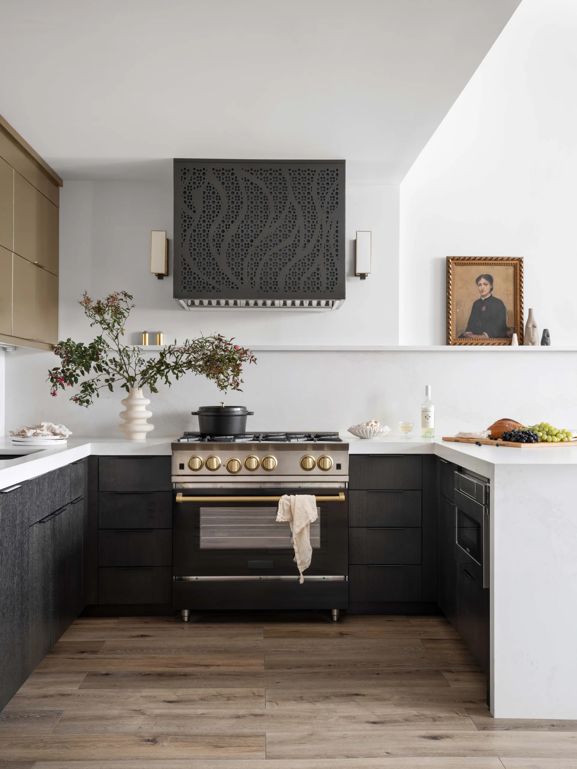 A modern kitchen features dark wood lower cabinets, white stone countertops, and a custom black perforated range hood above a professional range.