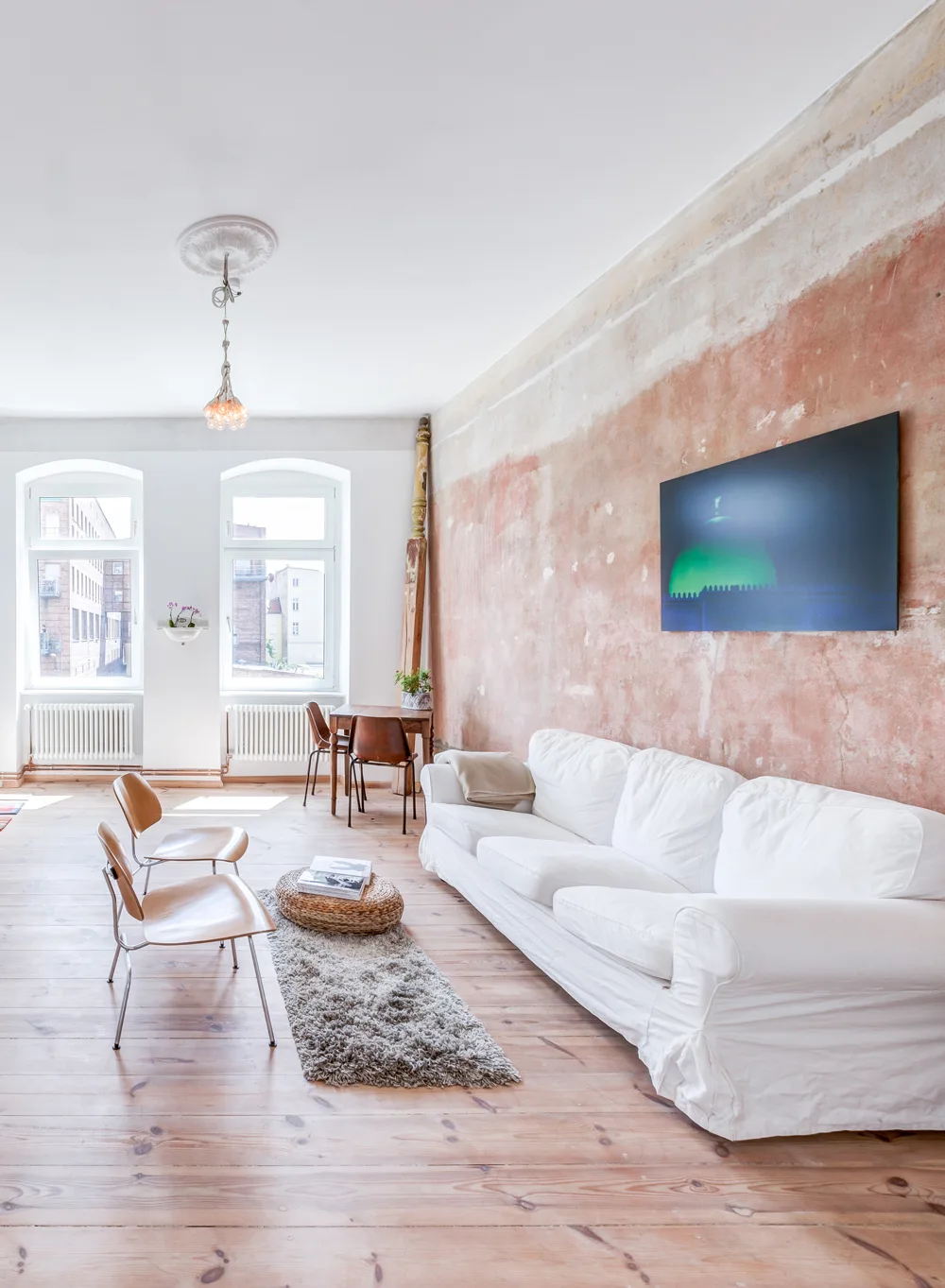 A living room features a white slipcovered sofa against a weathered terracotta plaster wall, light wood flooring, and two arched windows.