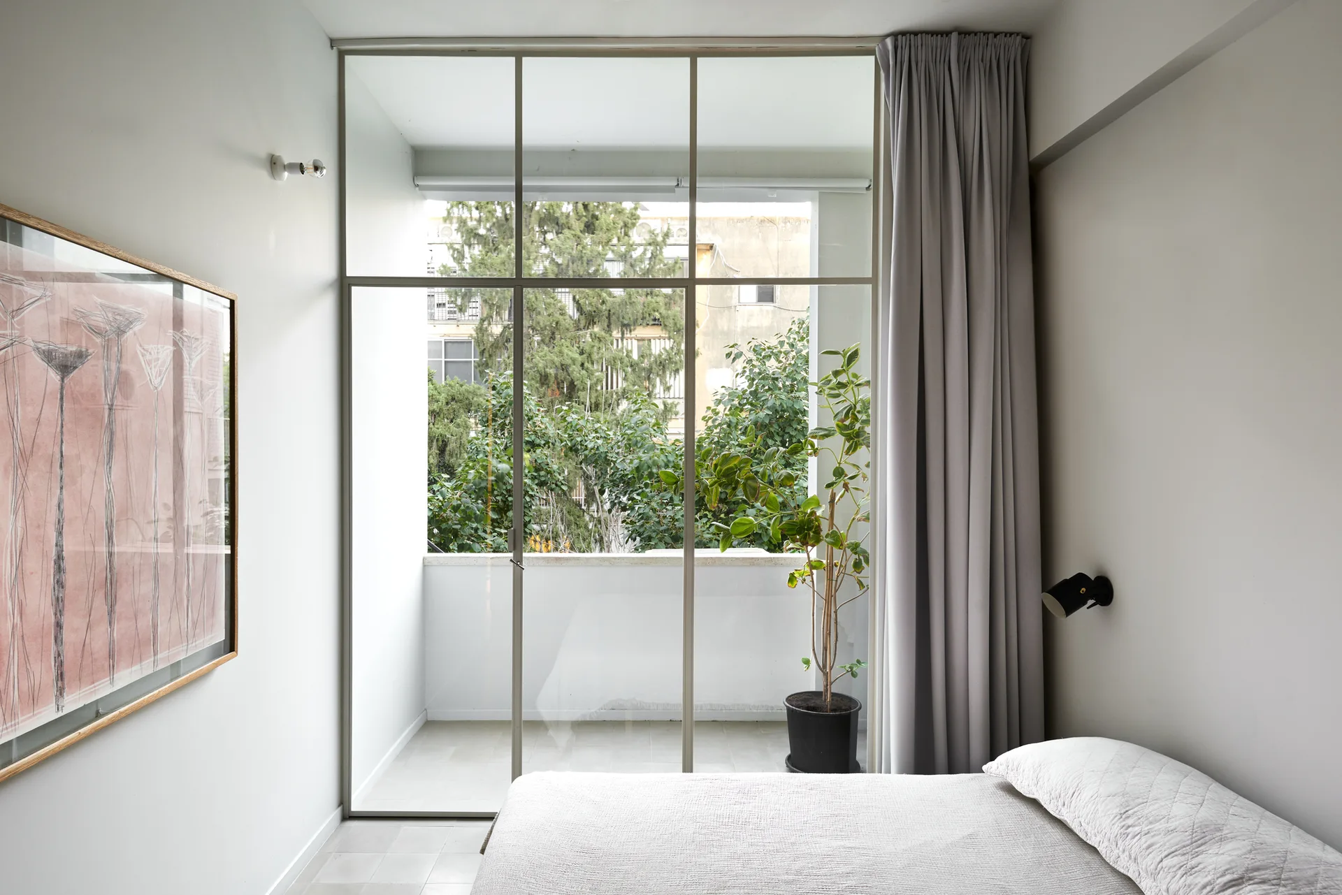 A minimalist bedroom featuring a low bed with white quilted bedding, a large framed art piece, and tall glass doors opening to a balcony.