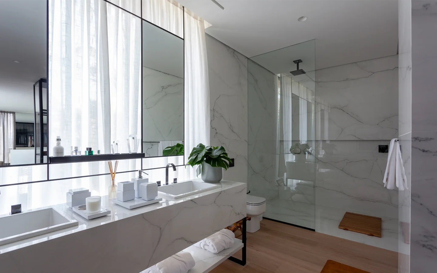 A modern bathroom features a white veined marble floating double vanity, black fixtures, sheer white curtains, and a glass-enclosed shower on wood flooring.