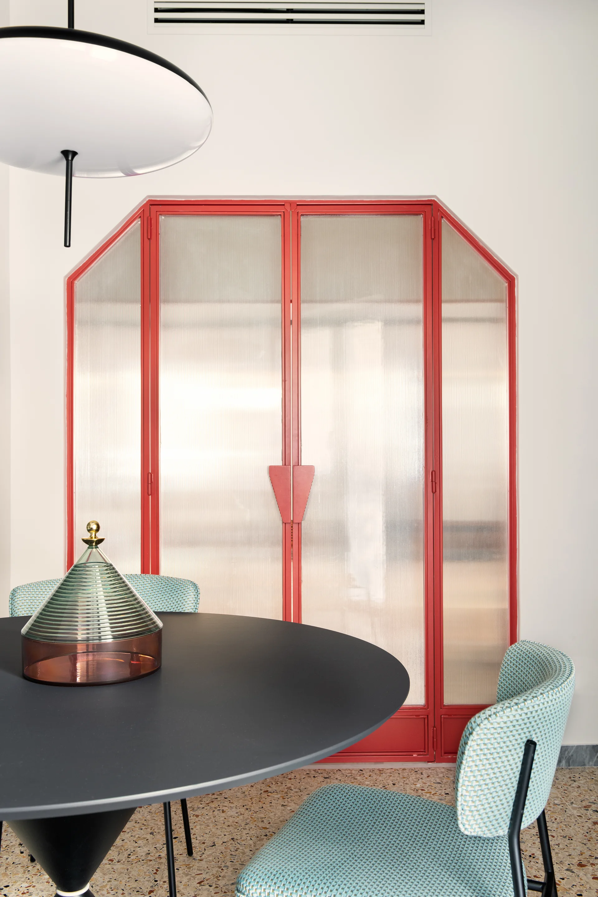 A modern dining area features a black round table and textured turquoise chairs set against a red-framed fluted glass partition door on terrazzo flooring.
