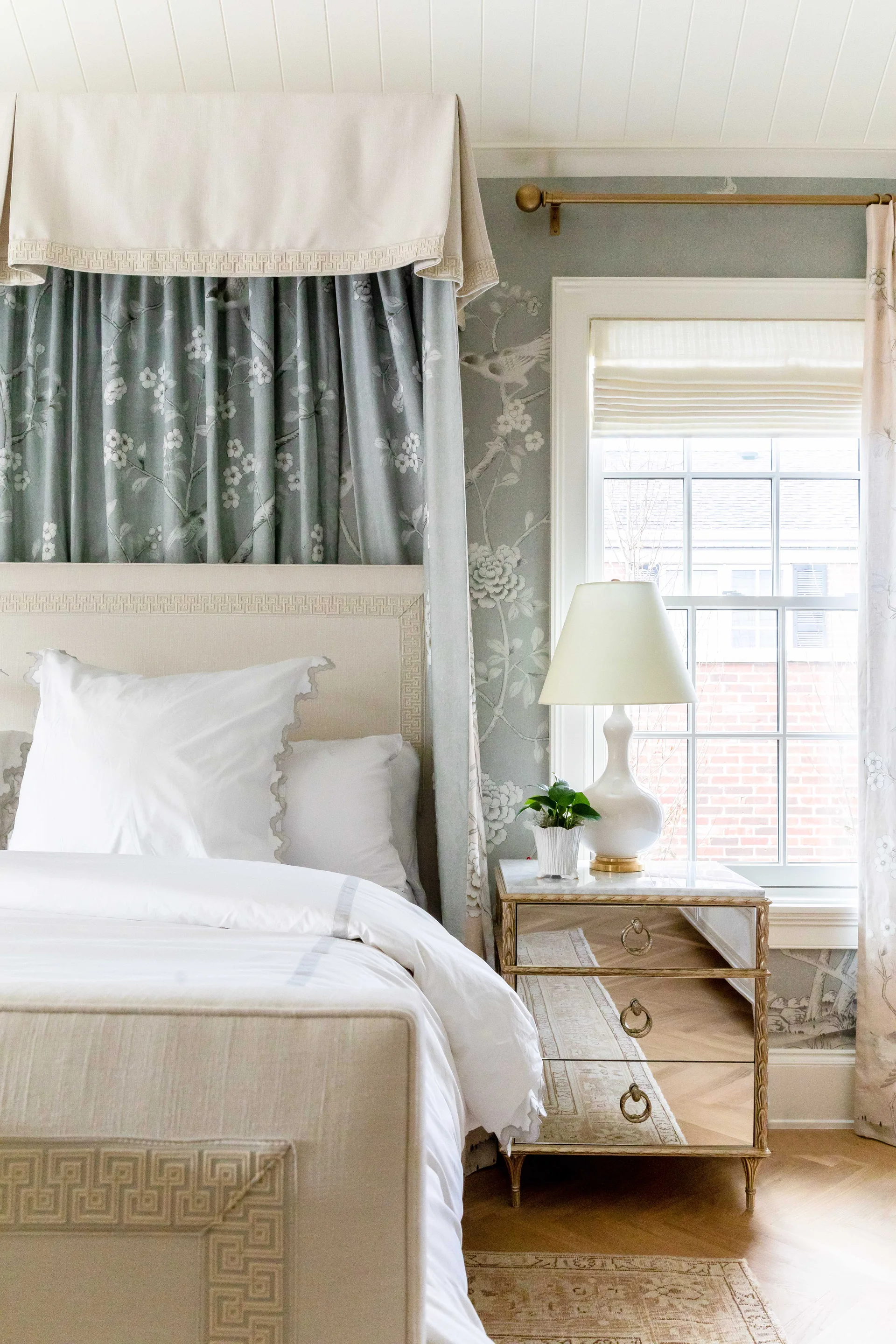 A canopy bed with blue floral drapery stands next to a mirrored three-drawer nightstand and a window in a bedroom with patterned wallpaper.