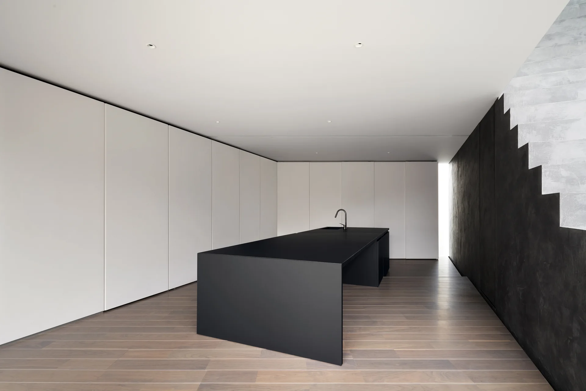 A minimalist kitchen features a matte black island with a faucet, set against seamless white cabinets, wood plank flooring, and a dark textured staircase wall.