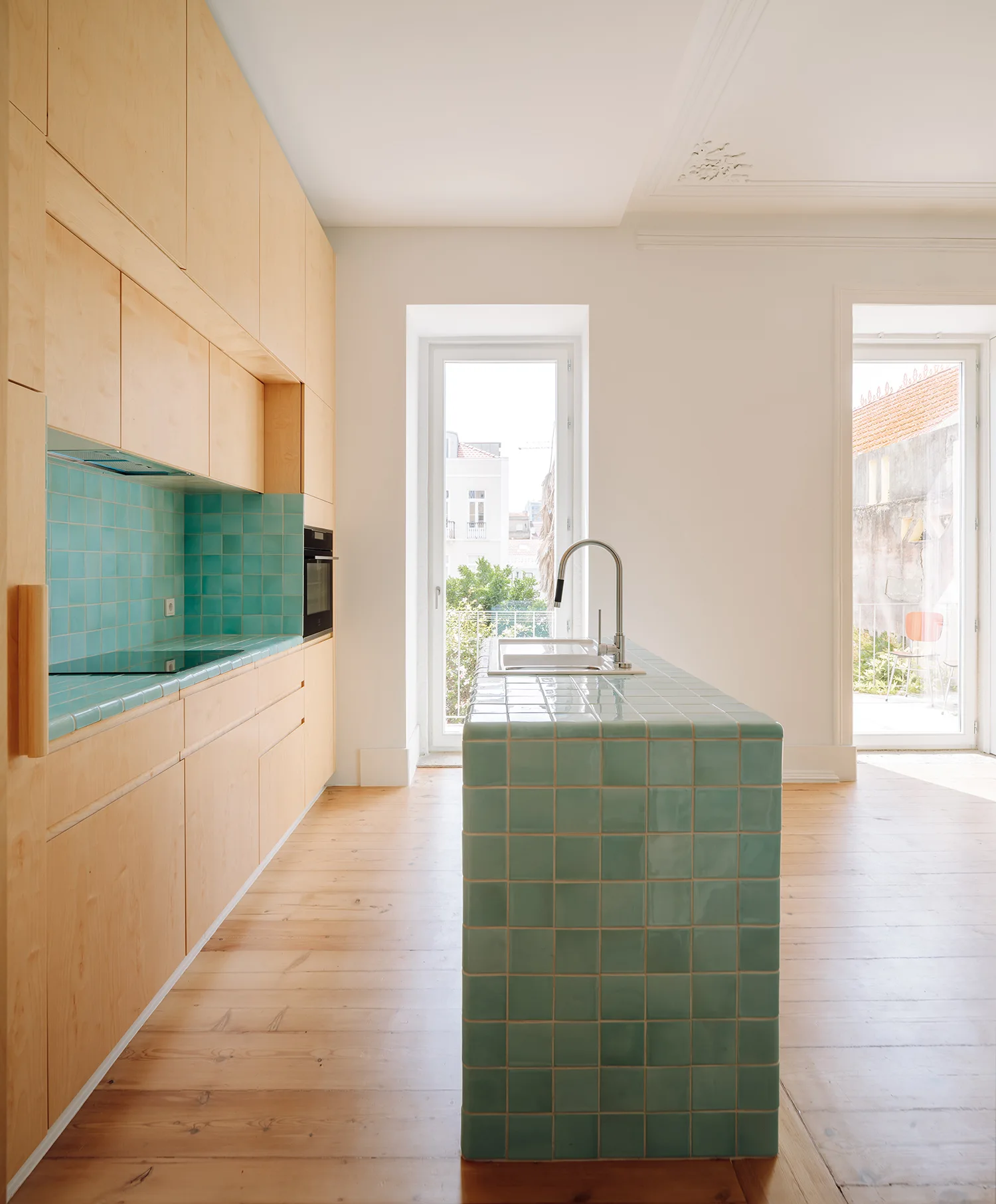 A minimalist kitchen features light wood cabinets alongside a central island and a matching backsplash covered in square green ceramic tiles.