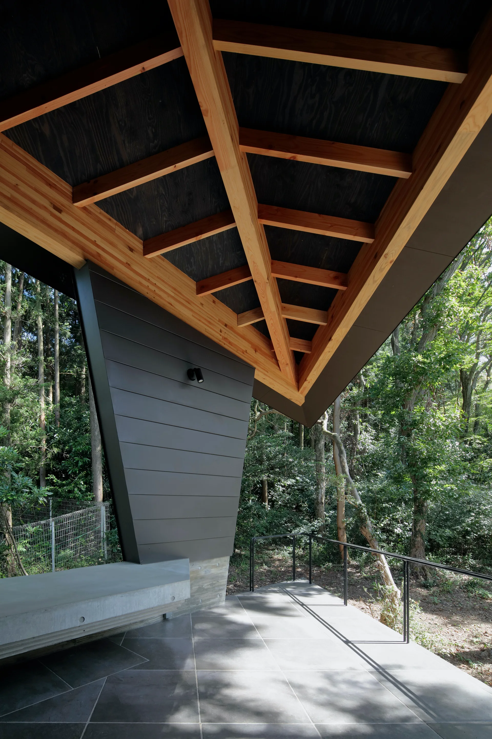 A modern covered patio features a natural timber rafter ceiling, a smooth concrete bench, dark horizontal wall paneling, and a gray tile floor.