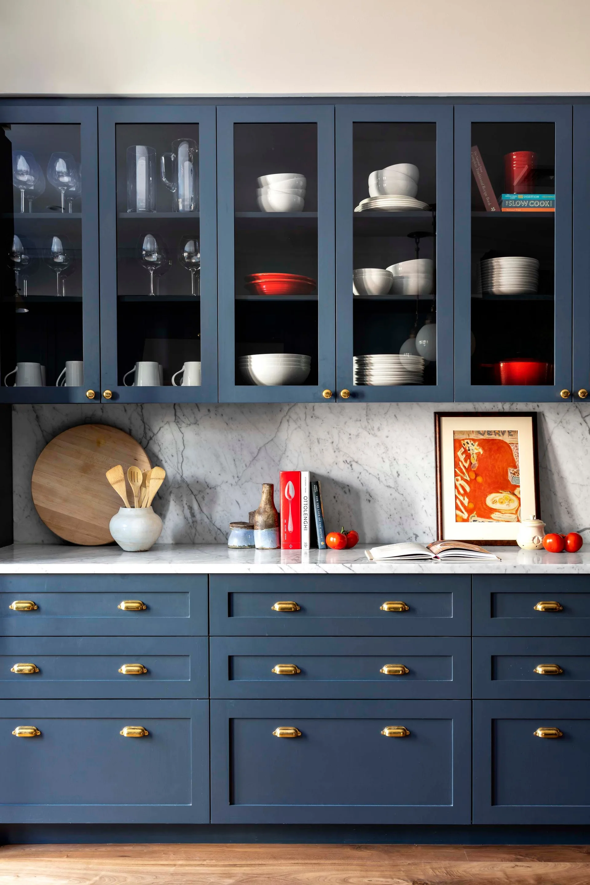 Blue shaker kitchen cabinets feature glass-front upper doors, a white marble backsplash, brass hardware, and stacked white dishware.