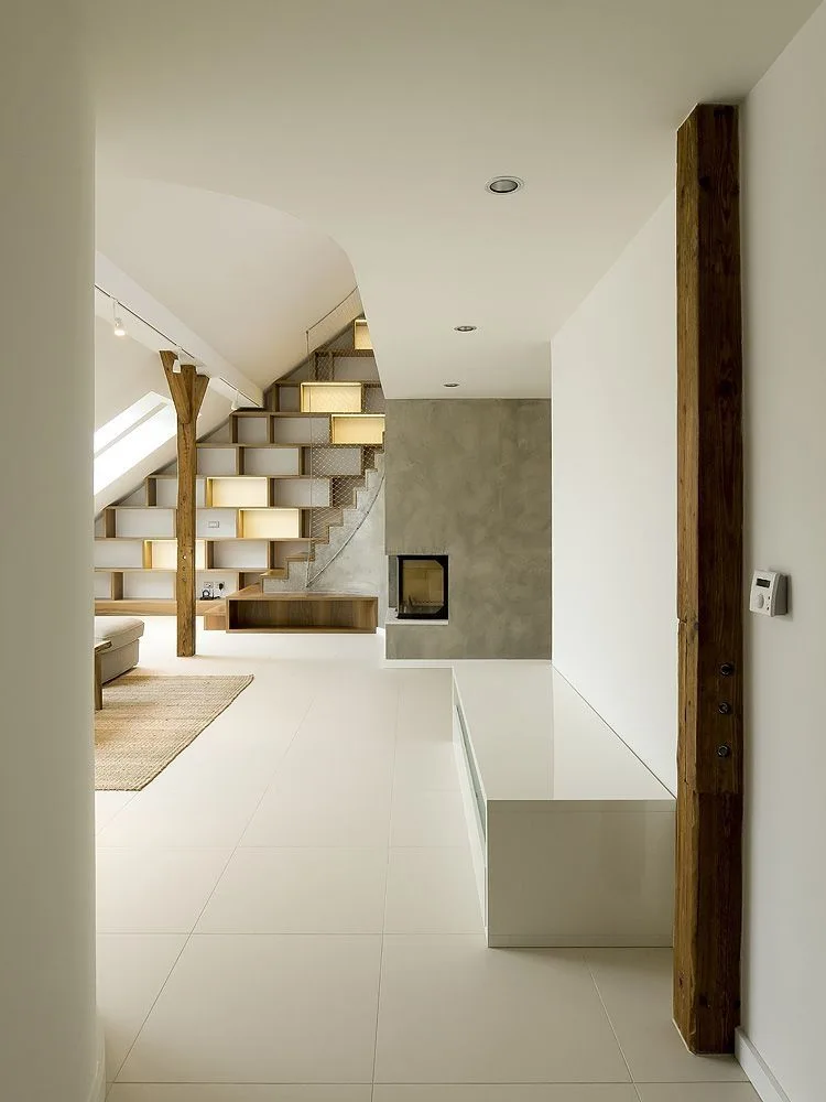 A modern loft interior features a staircase with built-in wood shelving, an exposed vertical beam, a concrete fireplace, and white tiled flooring.