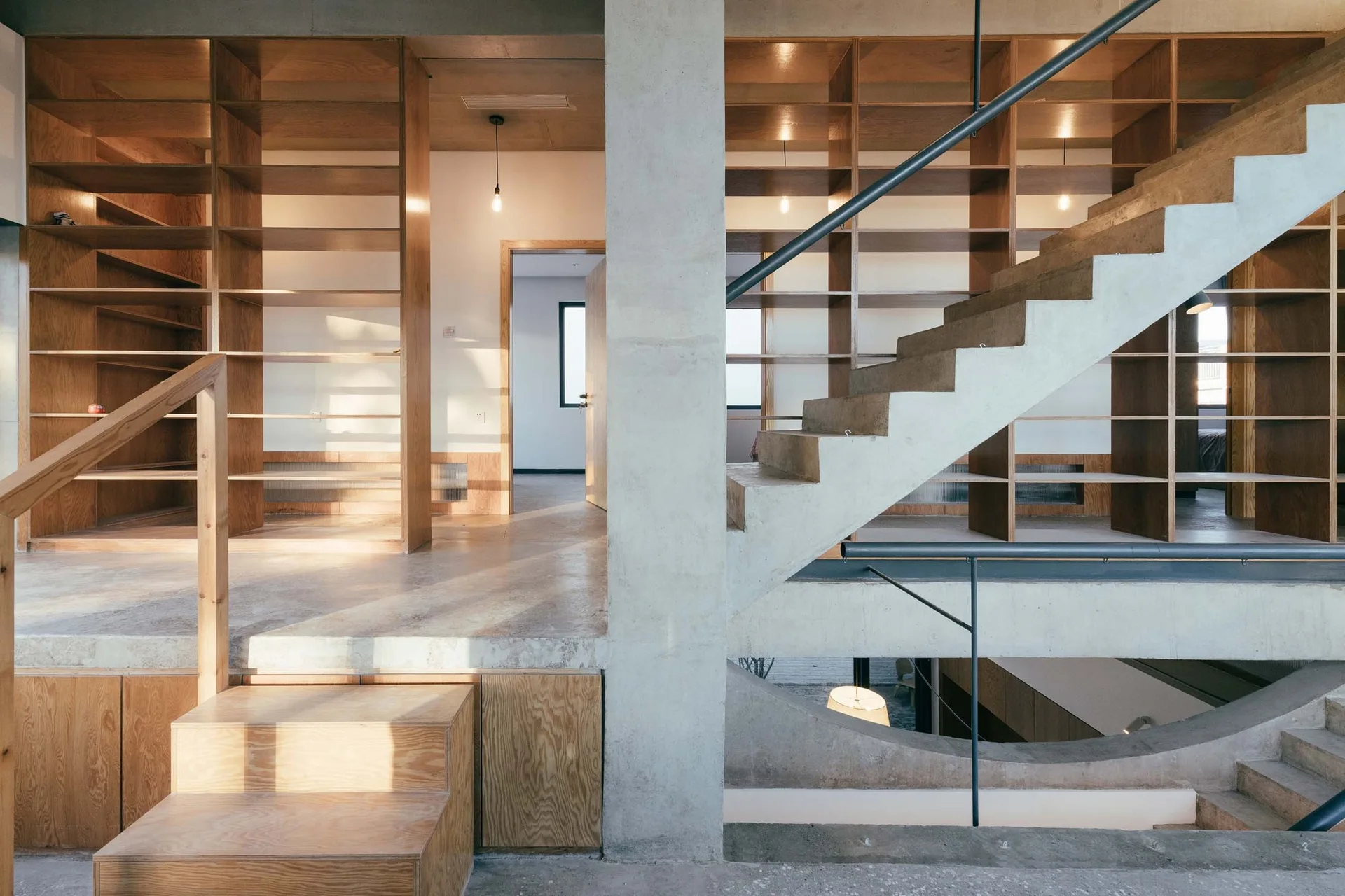 A multi-level interior features a raw concrete staircase, a central concrete column, and floor-to-ceiling built-in wooden shelving units.