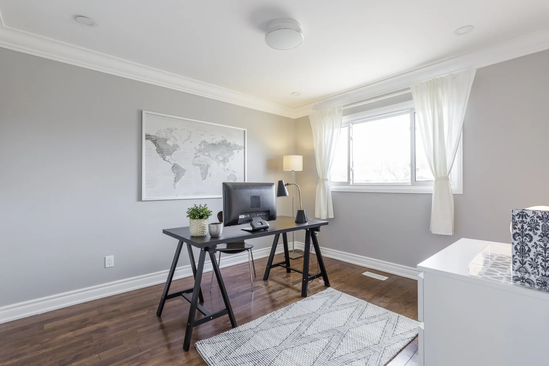 A minimalist home office featuring a black trestle desk, grey walls, dark wood floors, and a framed world map.