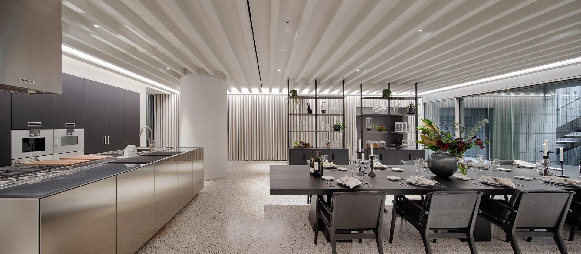 A modern open-concept kitchen and dining area with linear white ceiling baffles, a stainless steel island, and a dark wood table set for a meal.