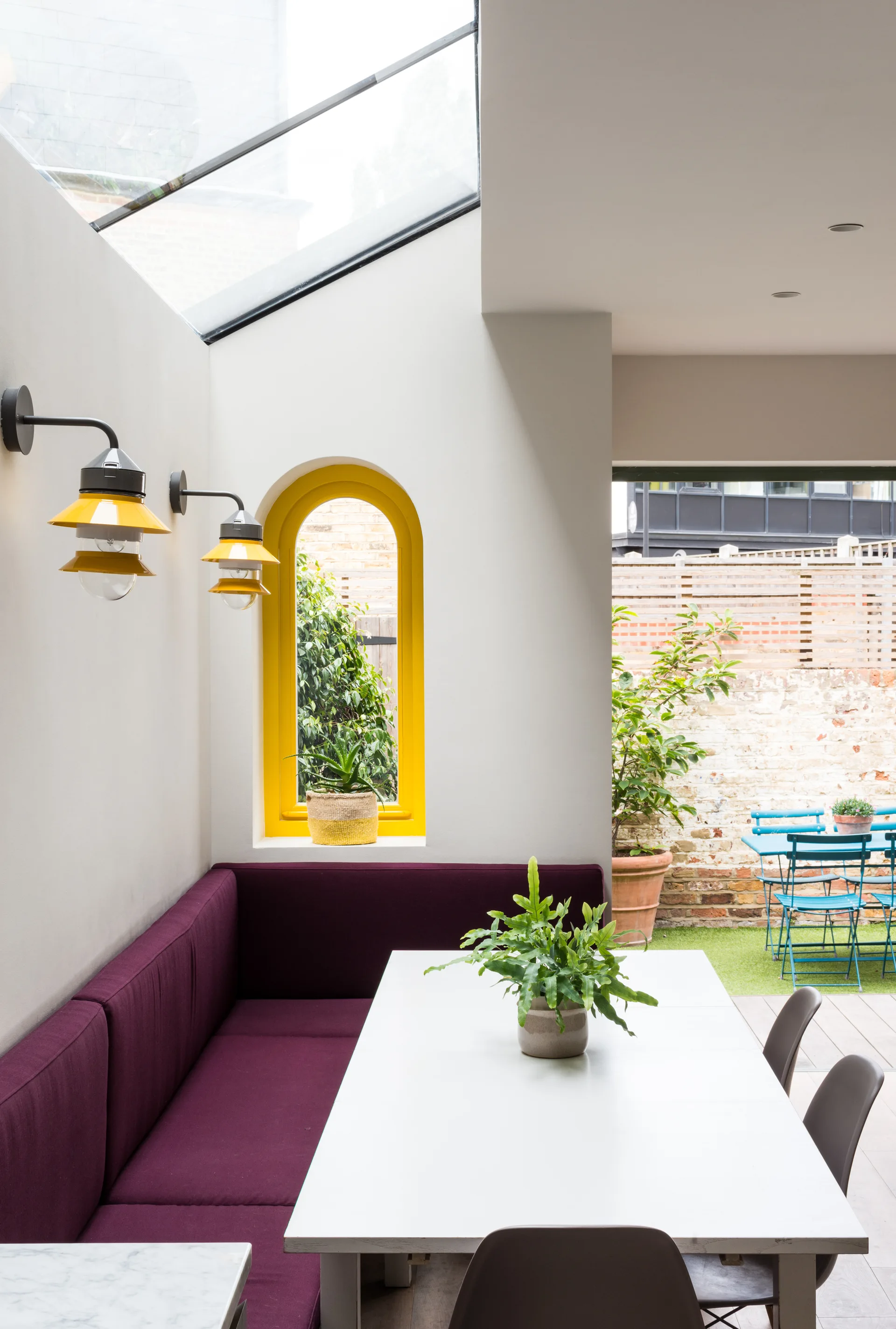 A bright dining area features a plum-colored built-in booth, a white table, two yellow wall lights, and an arched yellow window under a skylight.