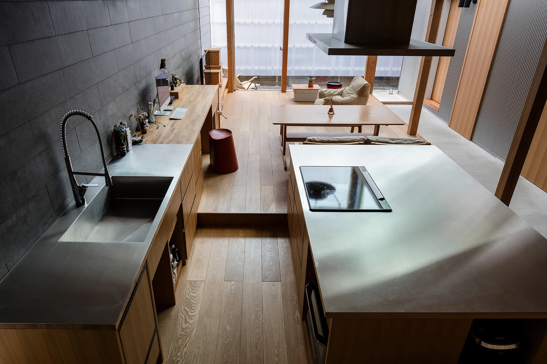 A high-angle view of a minimalist kitchen featuring a stainless steel island, wooden cabinetry, and a stepped wood floor leading to a dining area.