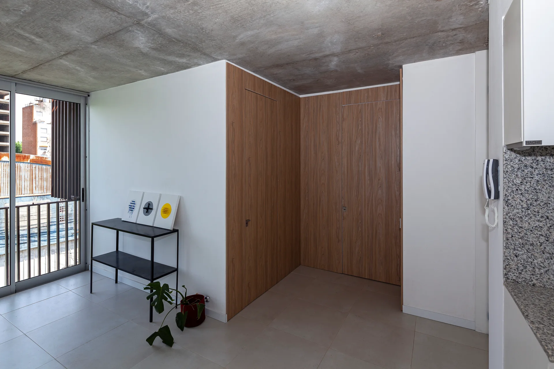 A minimalist apartment corner features an exposed concrete ceiling, large grey floor tiles, a black metal console table, and full-height wood-paneled closets.