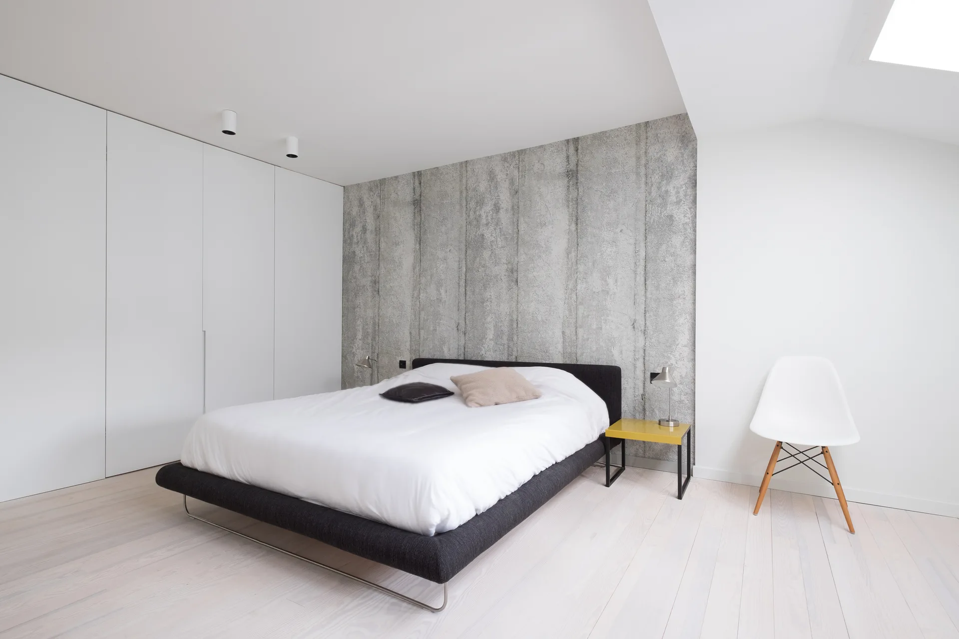 A modern minimalist bedroom features a grey platform bed against a textured concrete wall beside seamless white wardrobes.