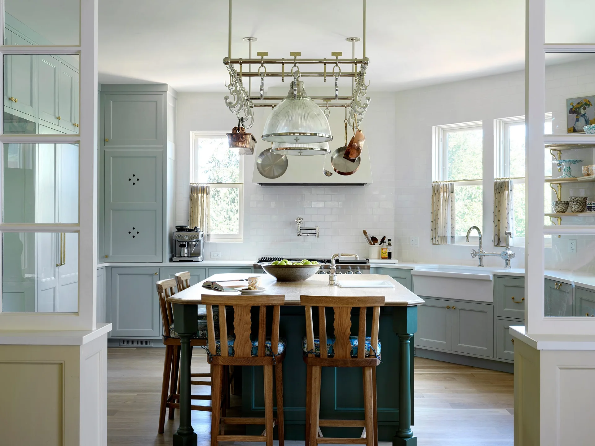 A bright kitchen features sage green cabinetry, a dark green central island with wooden stools, a white subway tile backsplash, and a hanging metal pot rack.