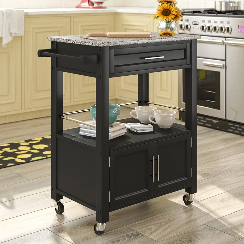 A black wooden kitchen cart on wheels features a granite top, a storage drawer, and a bottom cabinet on a light wood floor.