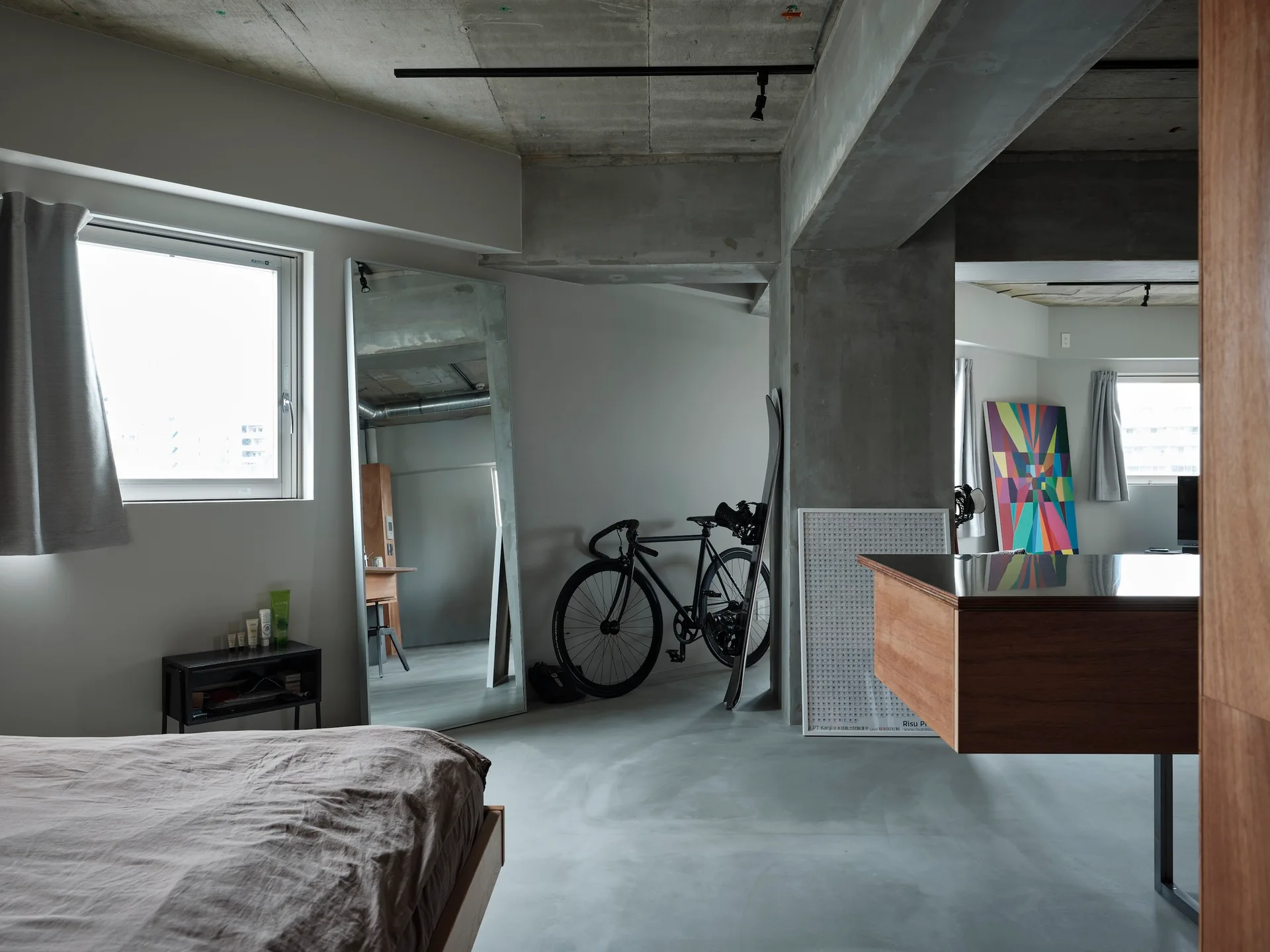 A minimalist bedroom featuring a raw concrete ceiling, a tall leaning floor mirror, a bed with gray bedding, and a black bicycle.