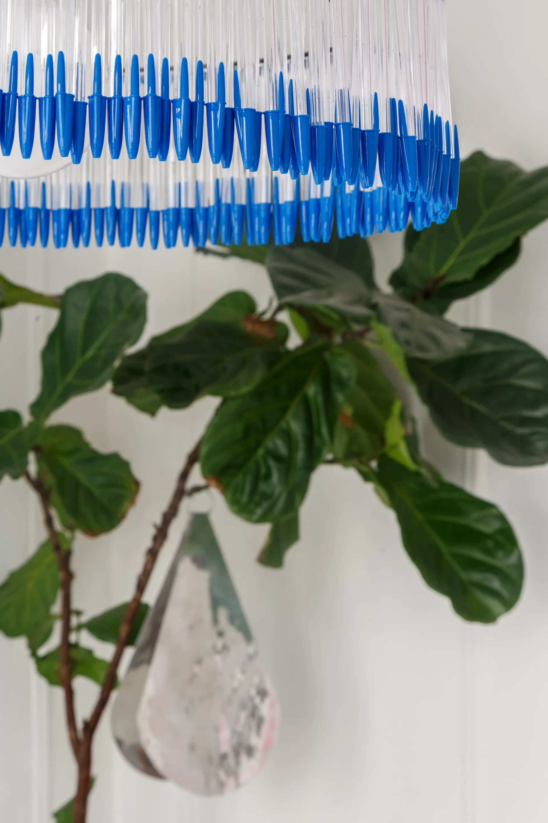 A circular pendant light fixture constructed from blue and clear plastic pens hangs in front of a green fiddle-leaf fig plant.