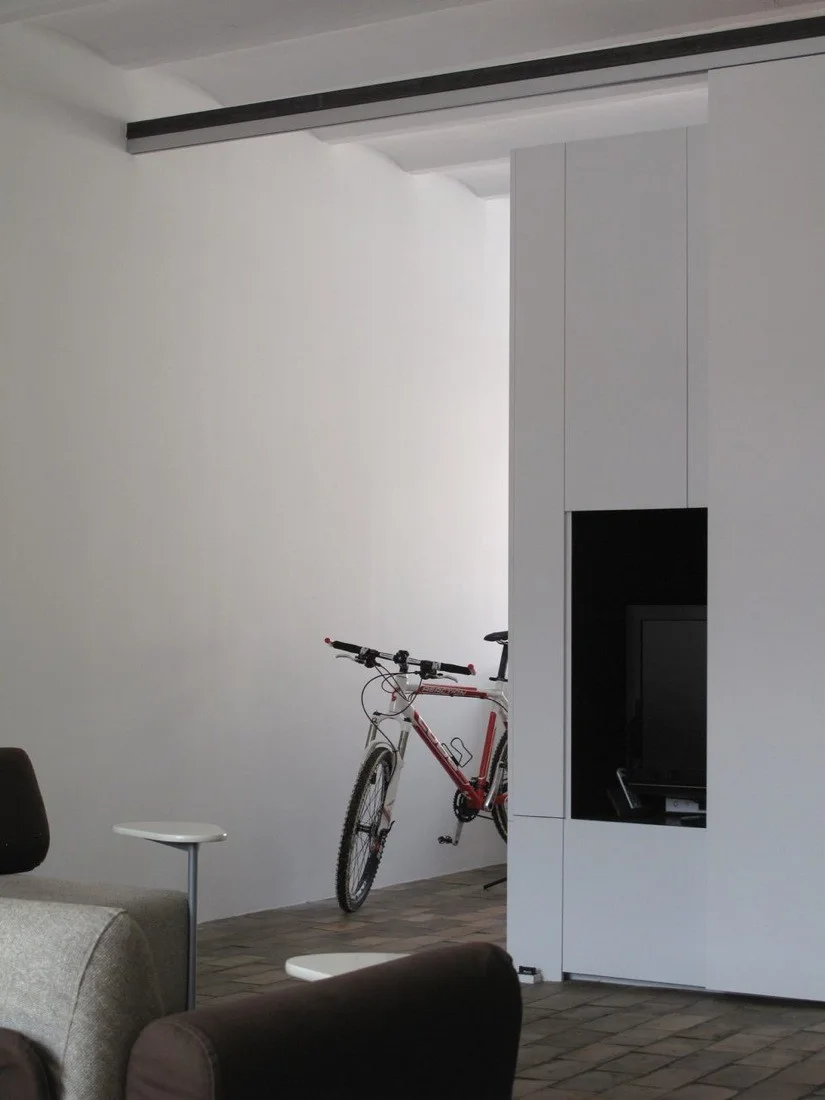 A modern living room features a floor-to-ceiling white cabinet with a recessed black TV niche, a bicycle against a white wall, and tile floors.