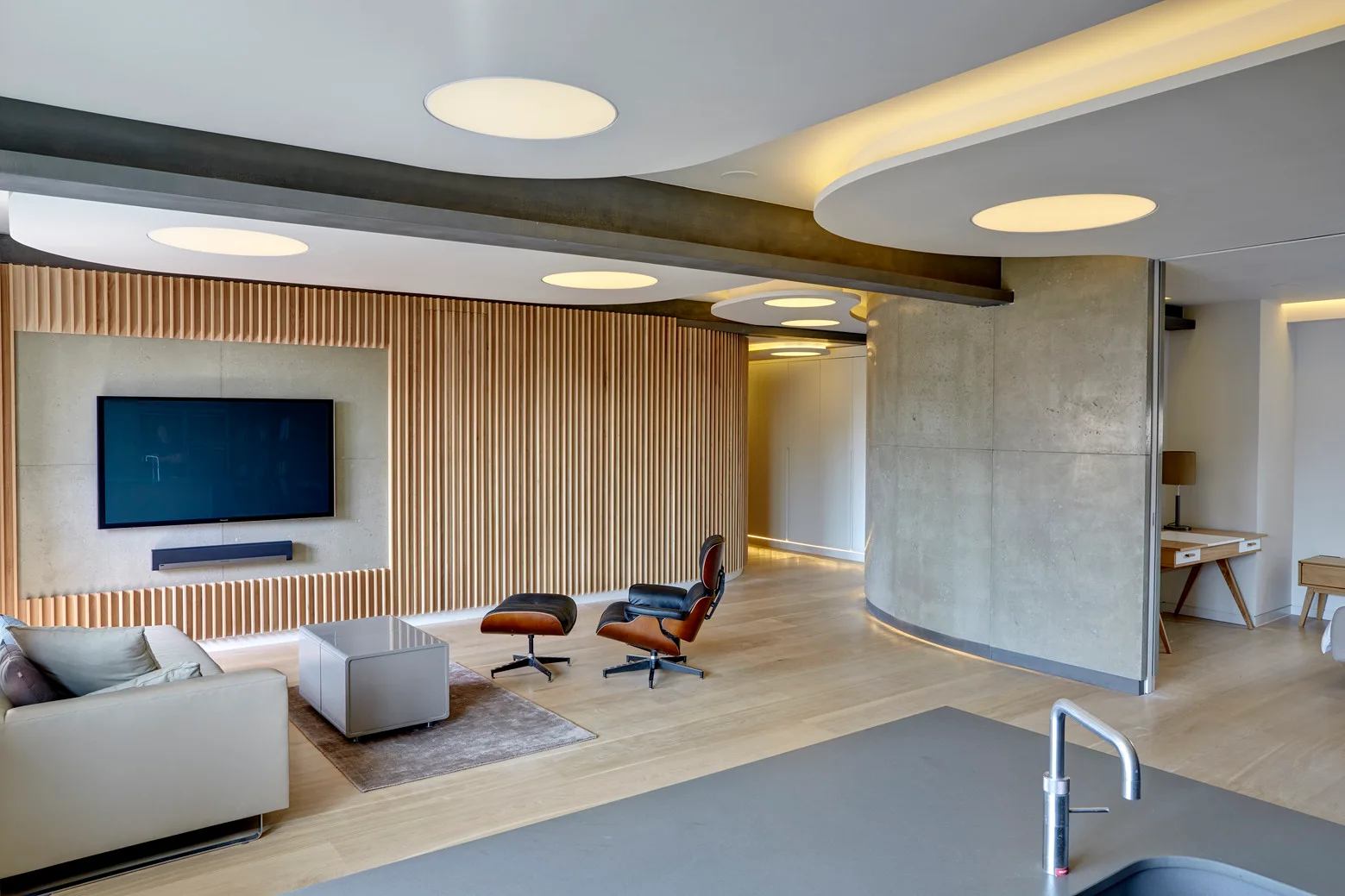 A modern open living room featuring a vertical wood slat wall, a curved concrete partition, and circular recessed ceiling lights.