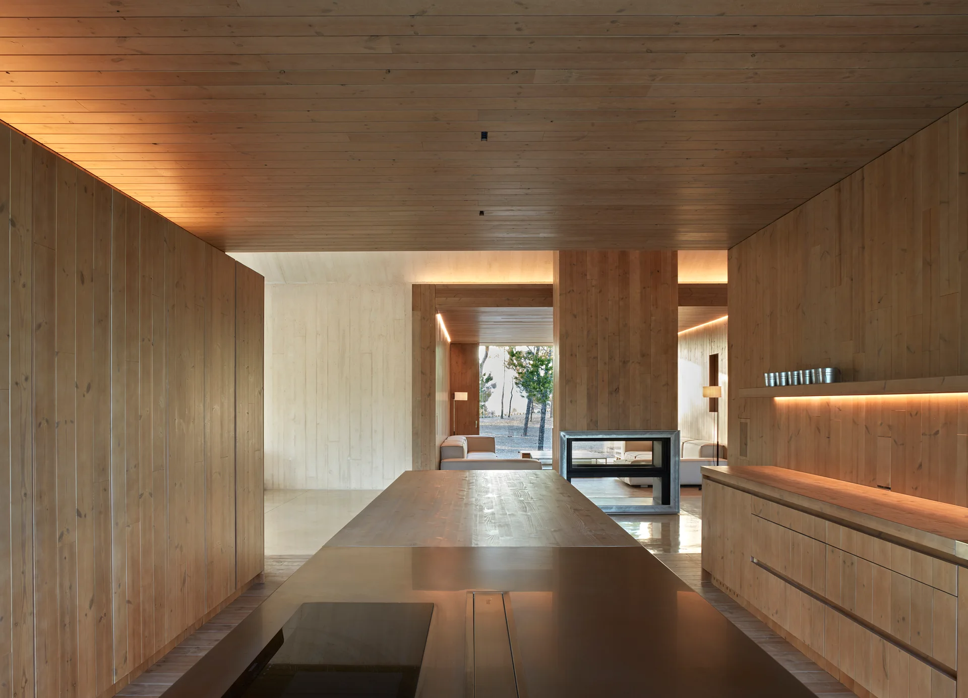 A minimalist kitchen featuring warm vertical wood plank cladding on the walls, ceiling, and island, looking toward an open living area.