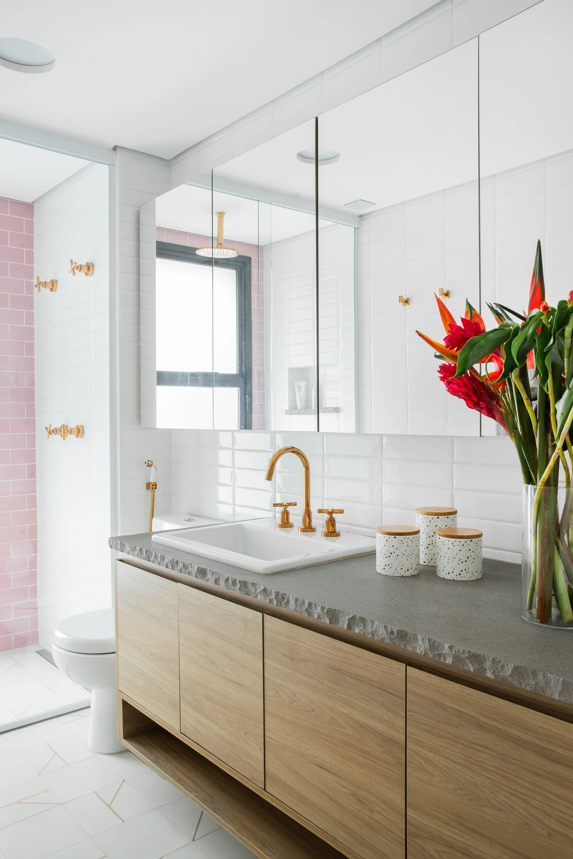A modern bathroom vanity features a light wood cabinet, a chiseled gray stone countertop, gold hardware, and a pink tiled shower in the background.