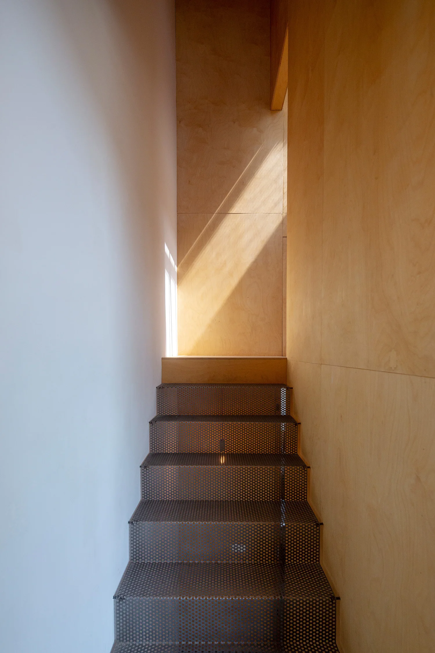 A narrow staircase features perforated metal steps set between a smooth white wall and natural plywood paneling under a beam of sunlight.