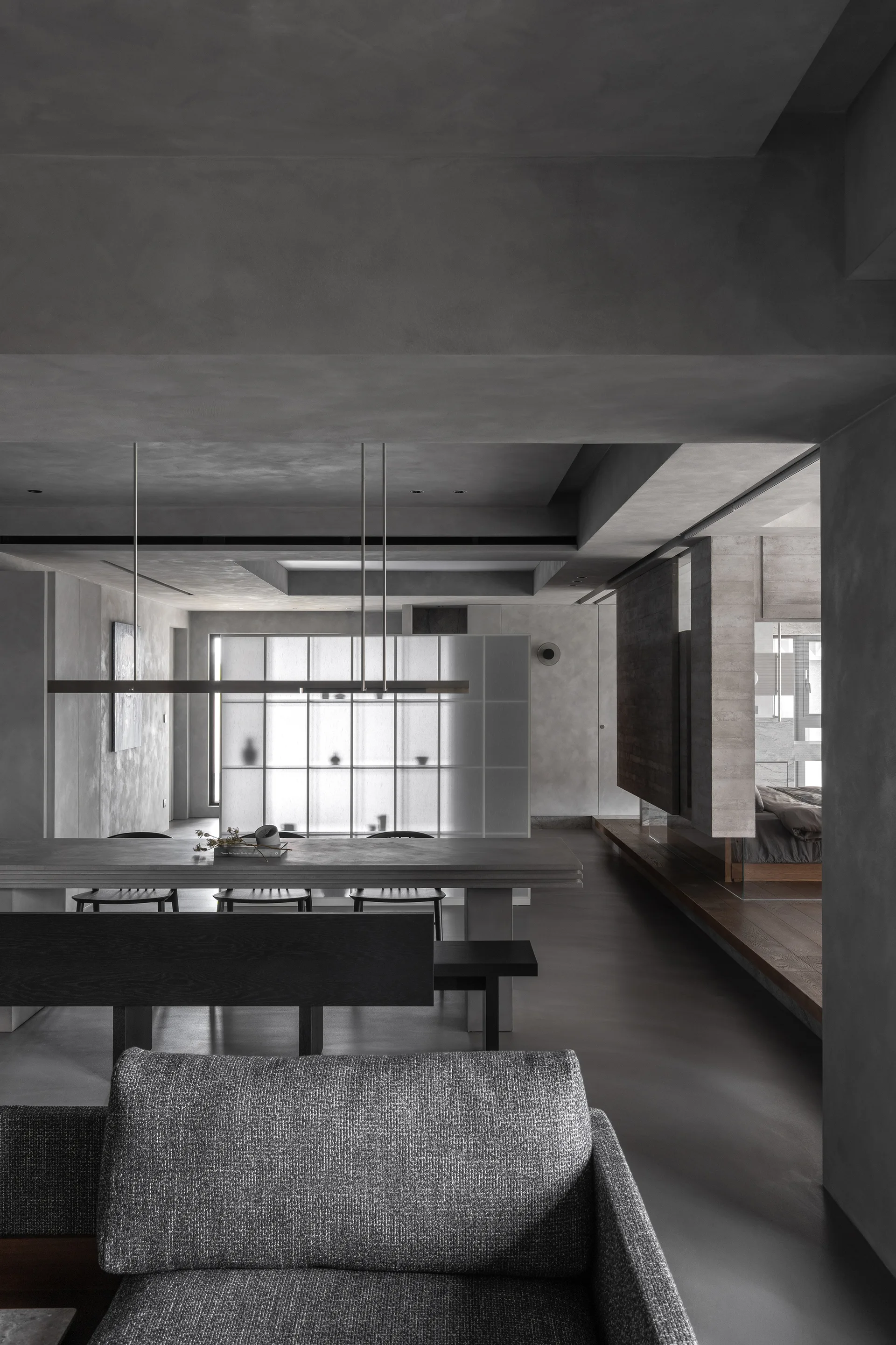 A grey minimalist dining space features a concrete dining table, black spindle chairs, a textured grey sofa, and a back-lit frosted glass grid partition.